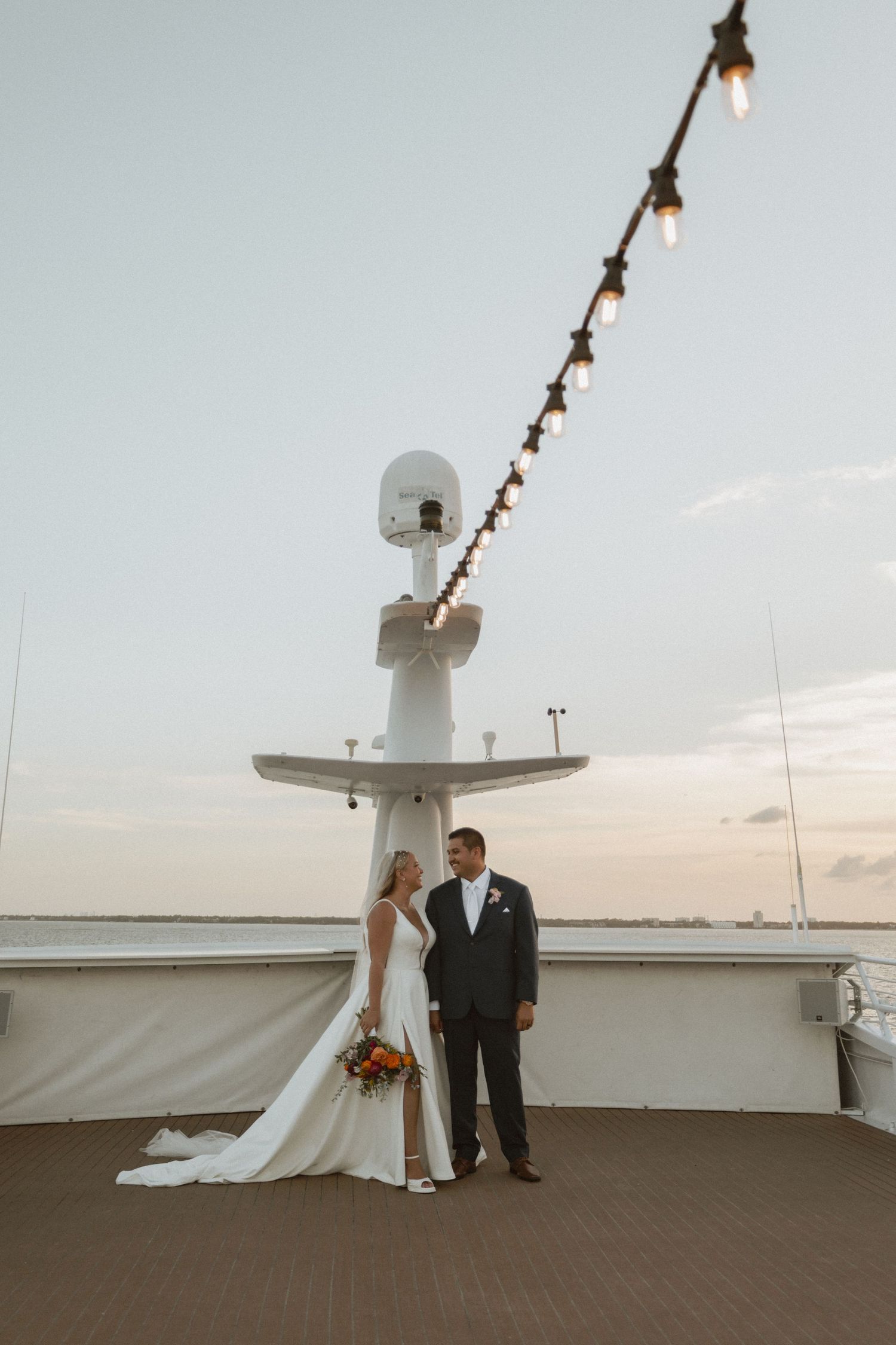 A couple poses under string lights on a boat deck at sunset with navigation equipment visible above.