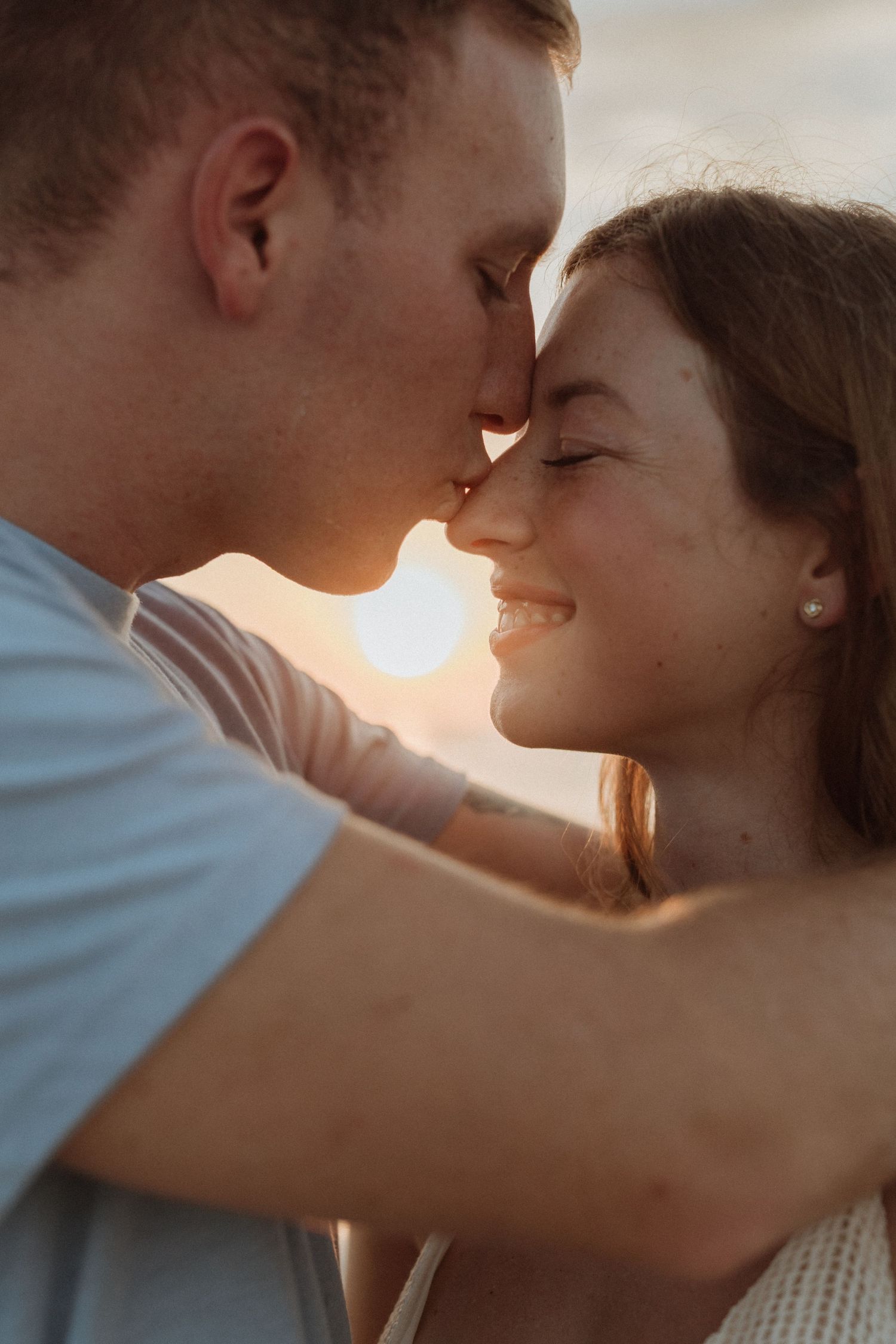 A couple shares a romantic sunset moment, touching foreheads and smiling tenderly on a beach.