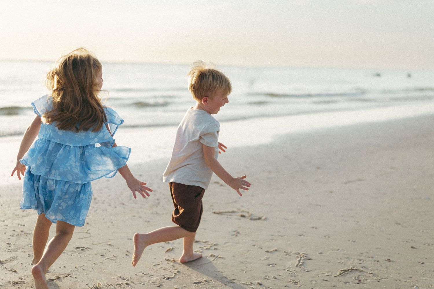 Two young children running barefoot on a sandy beach at sunset, girl in blue dress and boy in white shirt.
