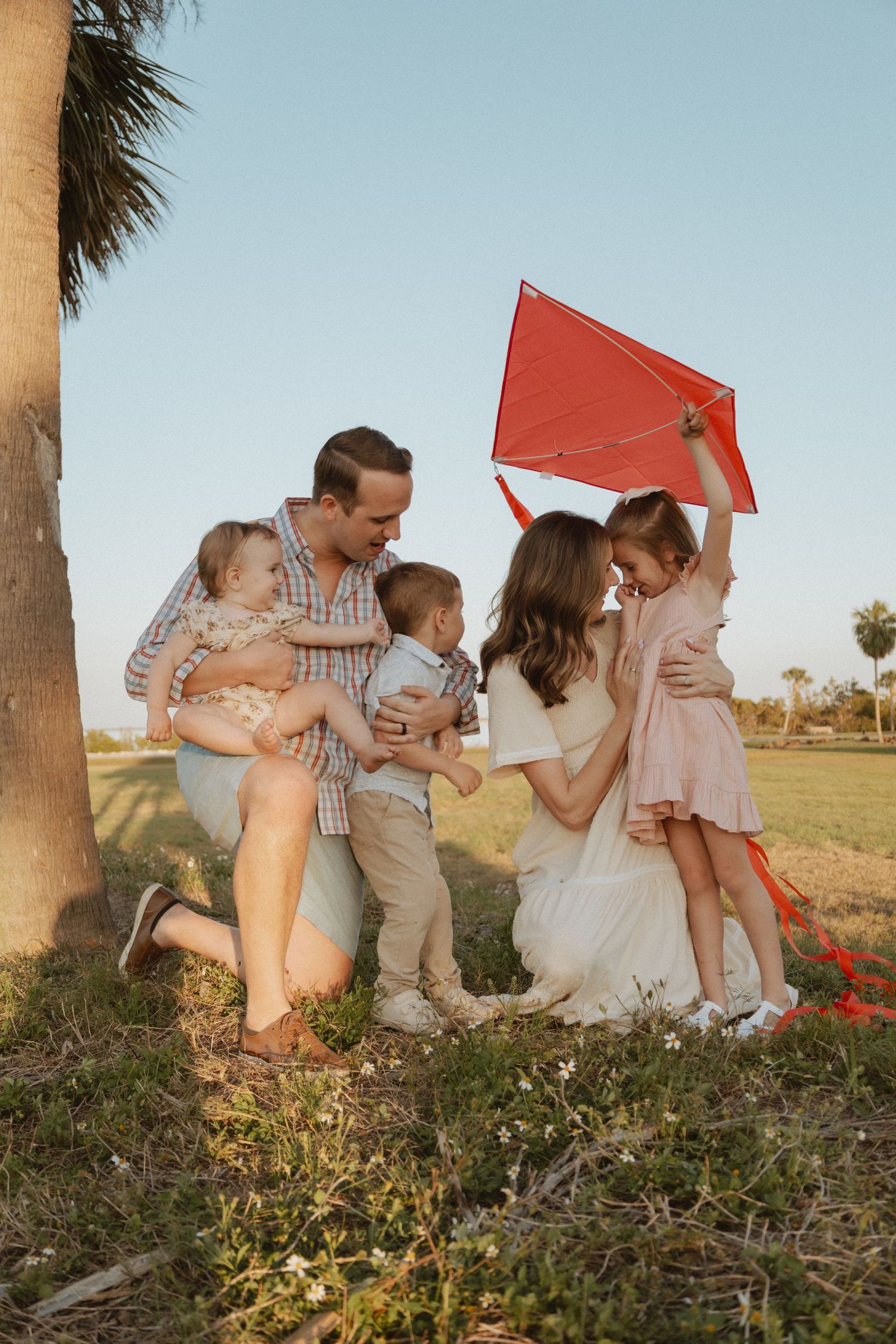 A warm family moment captured outdoors with a red kite and palm tree during golden hour.