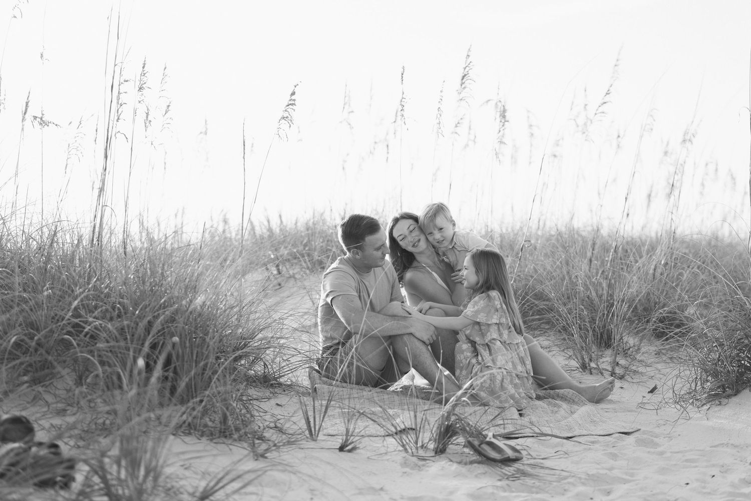 Black and white photo of a family sitting together on sandy beach dunes with tall sea oats grass in the background.