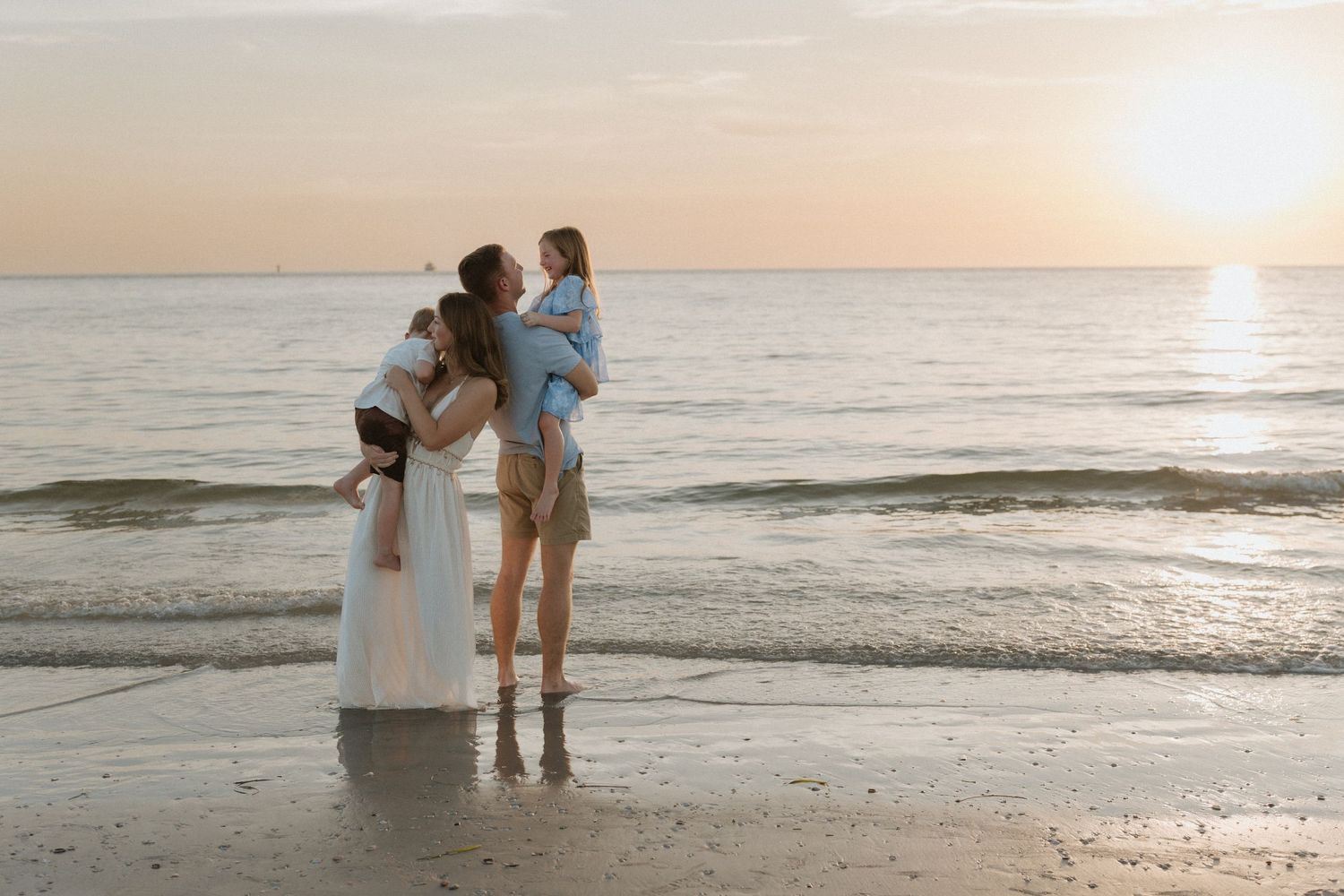 Family embracing at sunset on a beach, woman in white dress holding children near ocean waves.