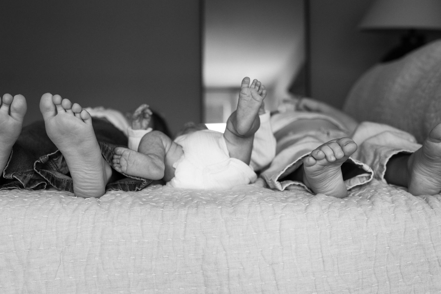 Black and white photo of newborn baby feet resting on a soft blanket beside adult feet.