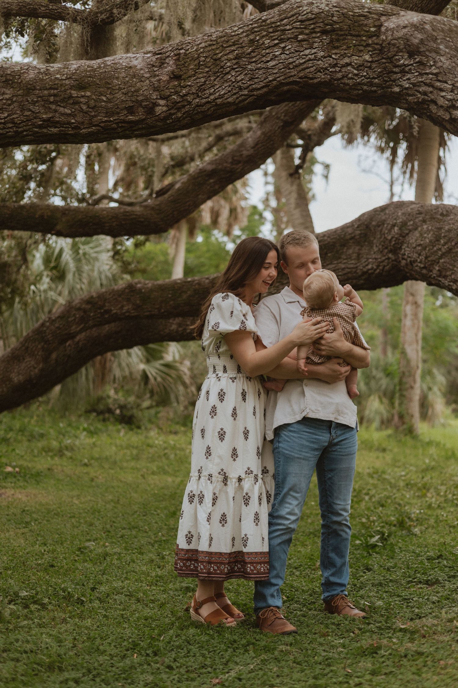 A couple shares an intimate moment beneath a large oak tree draped with Spanish moss in a natural outdoor setting.