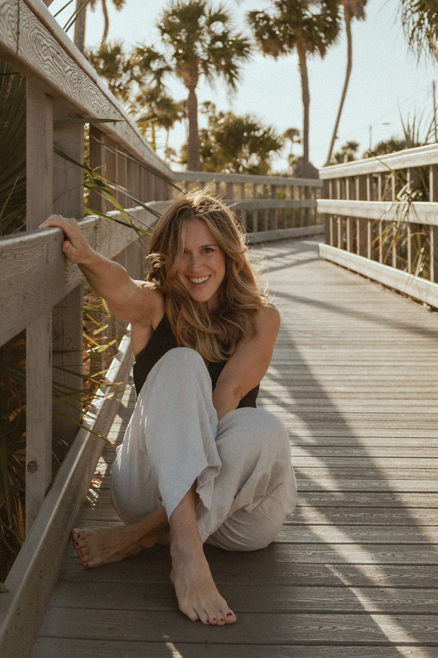Woman in casual attire relaxes on a wooden boardwalk with palm trees and sunlight creating a peaceful coastal scene.