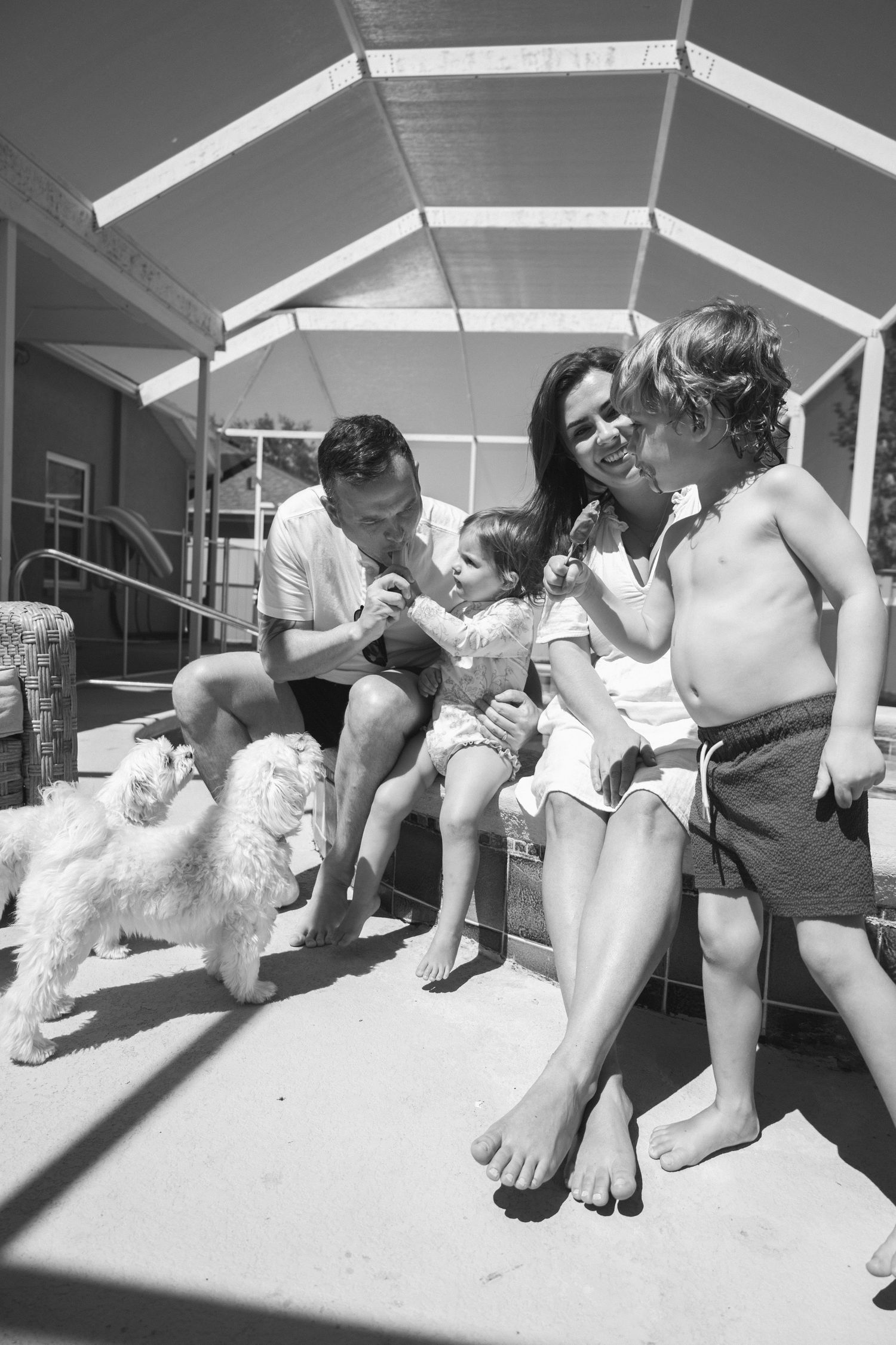 Black and white photo of a happy family with two children and a white fluffy dog relaxing under a poolside cabana.