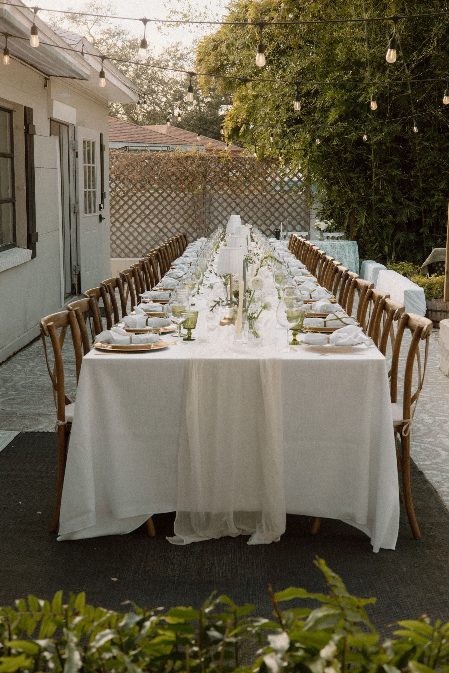 Elegant wedding reception table setup with white linens, dark wooden chairs, and romantic string lights overhead.