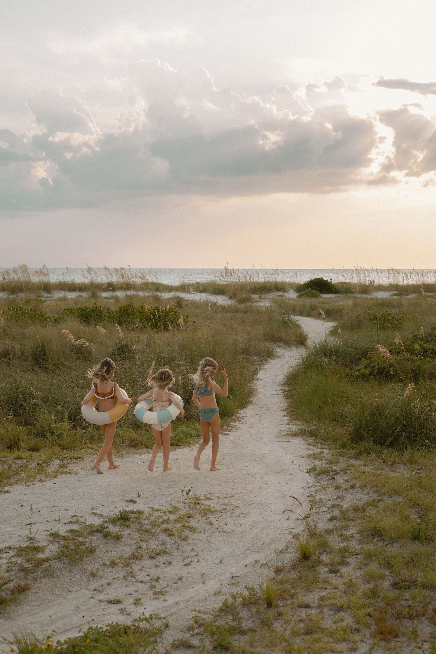 Friends walking along a sandy beach path surrounded by coastal vegetation at sunset.