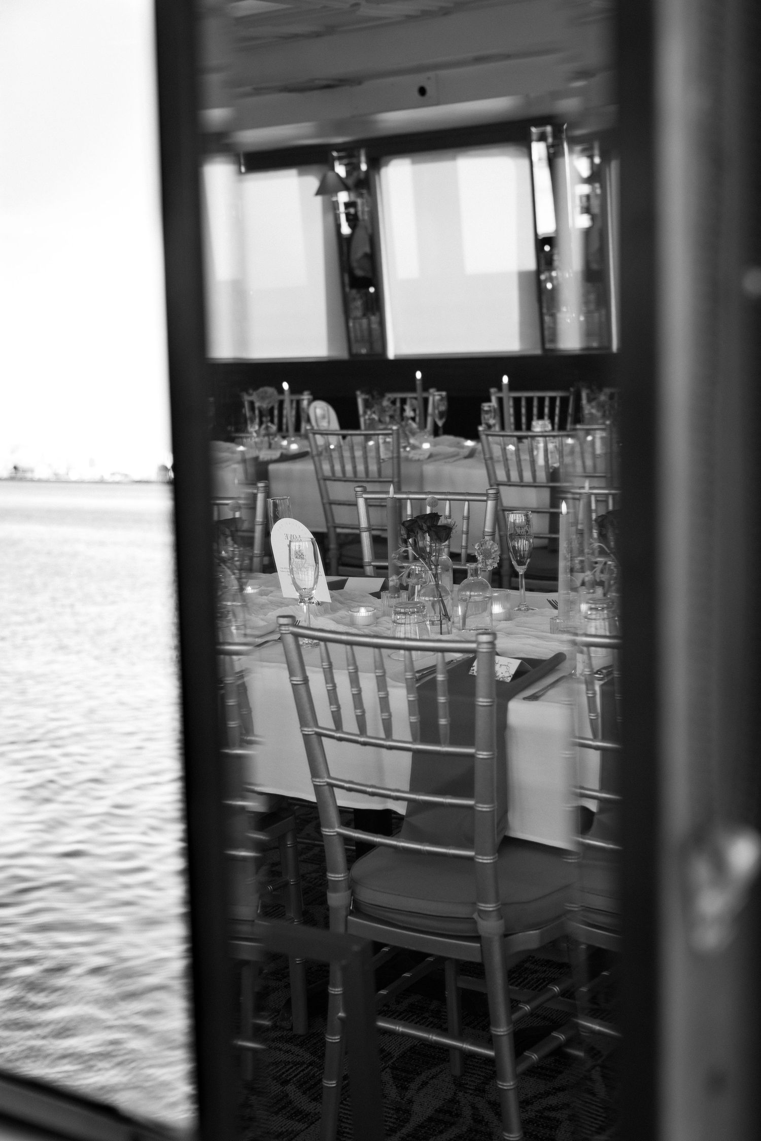 Black and white view through a window showing elegant table settings and chairs in a dining room.