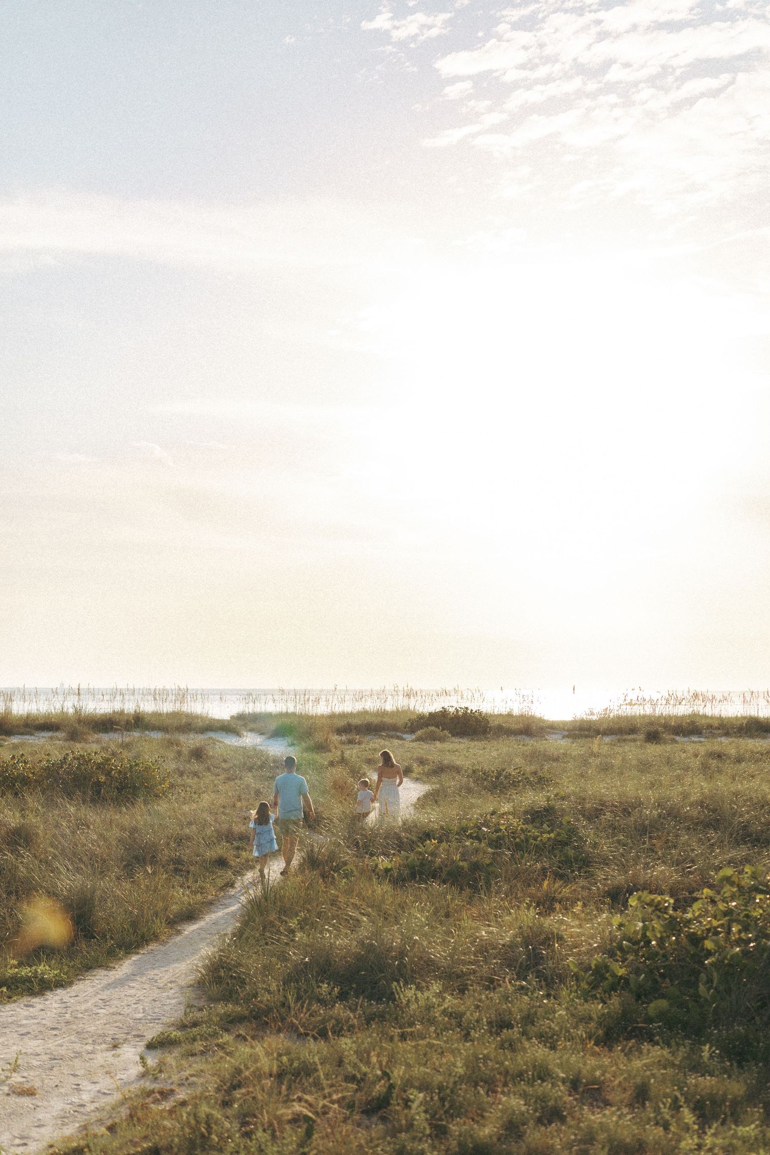 Family walking on a sunlit beach path through golden dunes toward the ocean at sunset.