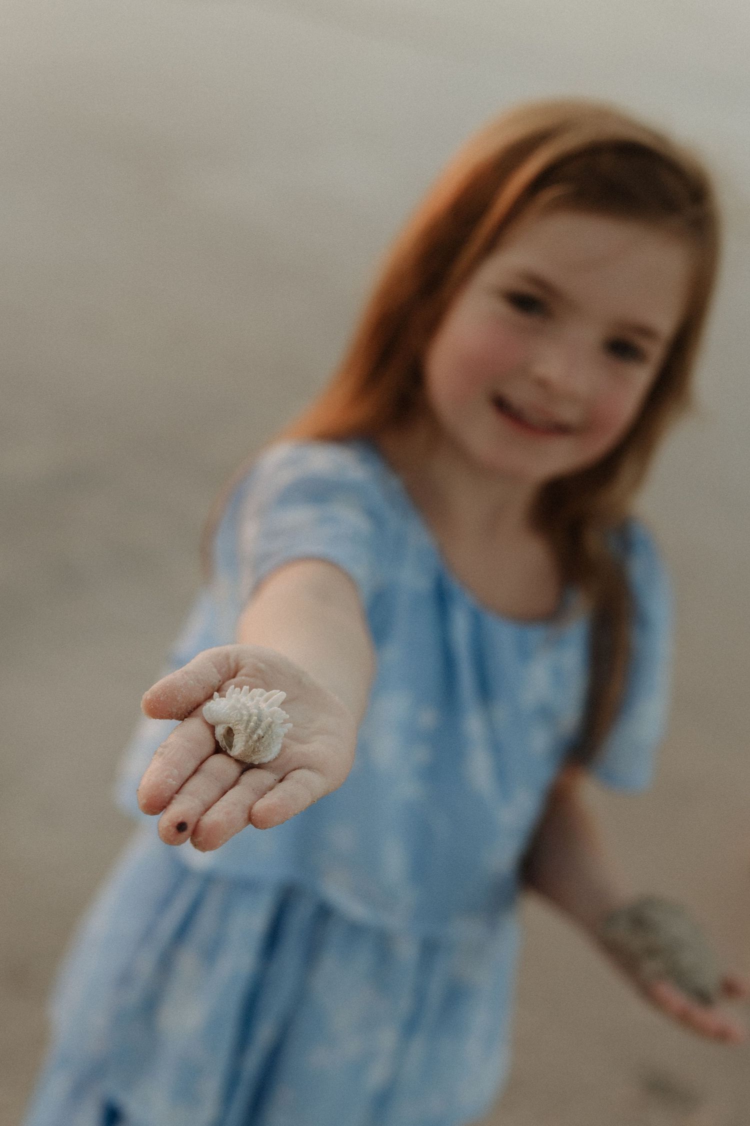 A smiling red-haired girl in a blue dress holds out a seashell on the beach.