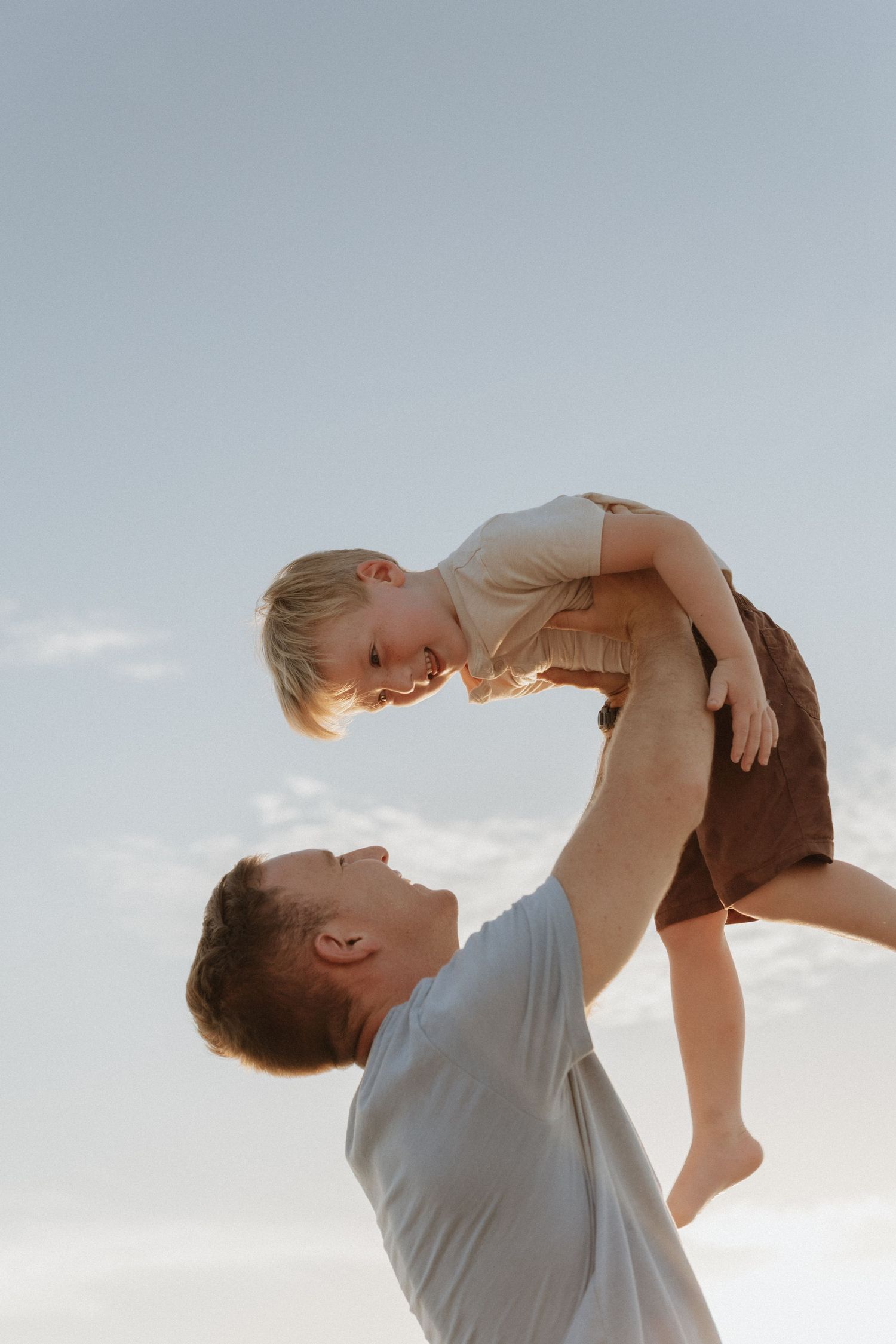A father joyfully lifts his young child high in the air against a clear sky, both smiling and sharing a playful moment.