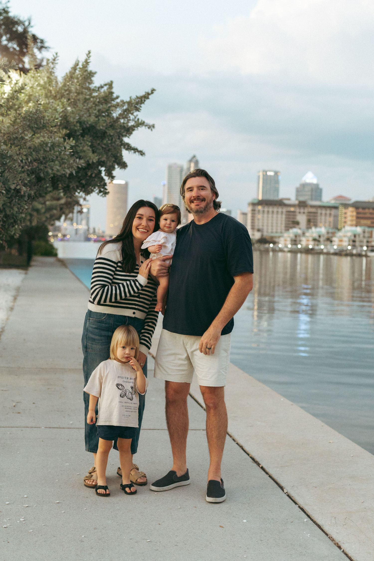 A family poses together for casual photos along a waterfront promenade with city buildings visible in the background.