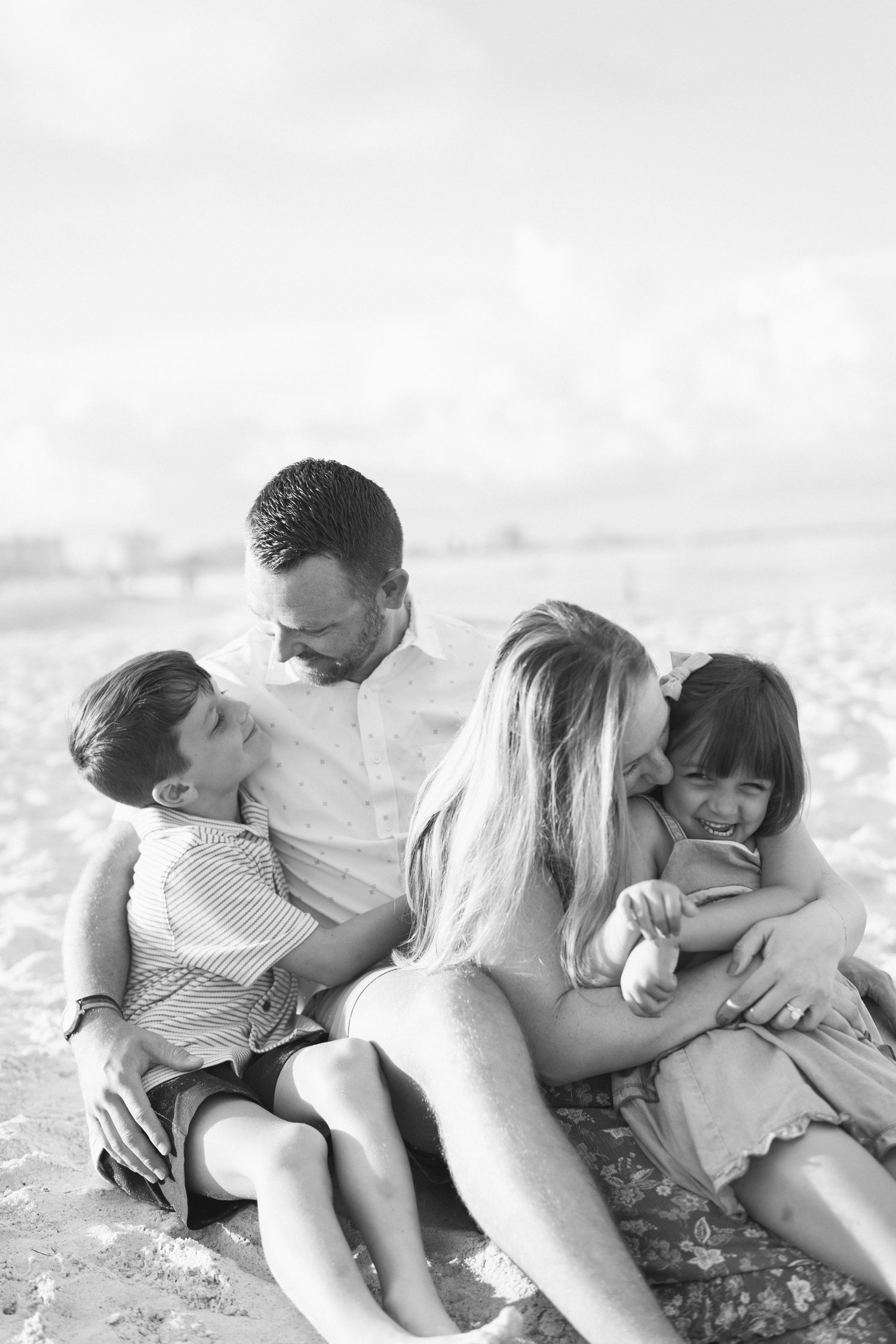 Black and white photo of a happy family of four sitting together on a sandy beach, sharing hugs and smiles.