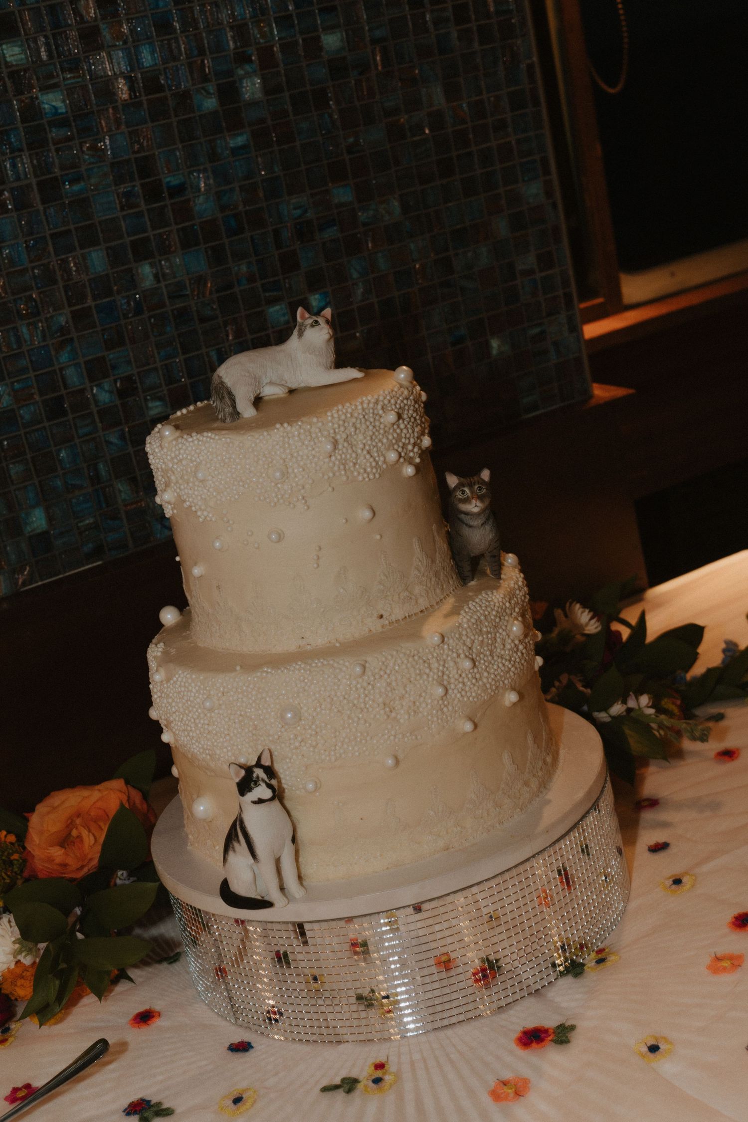 Three-tiered white wedding cake decorated with cat figurines on a sparkly stand.