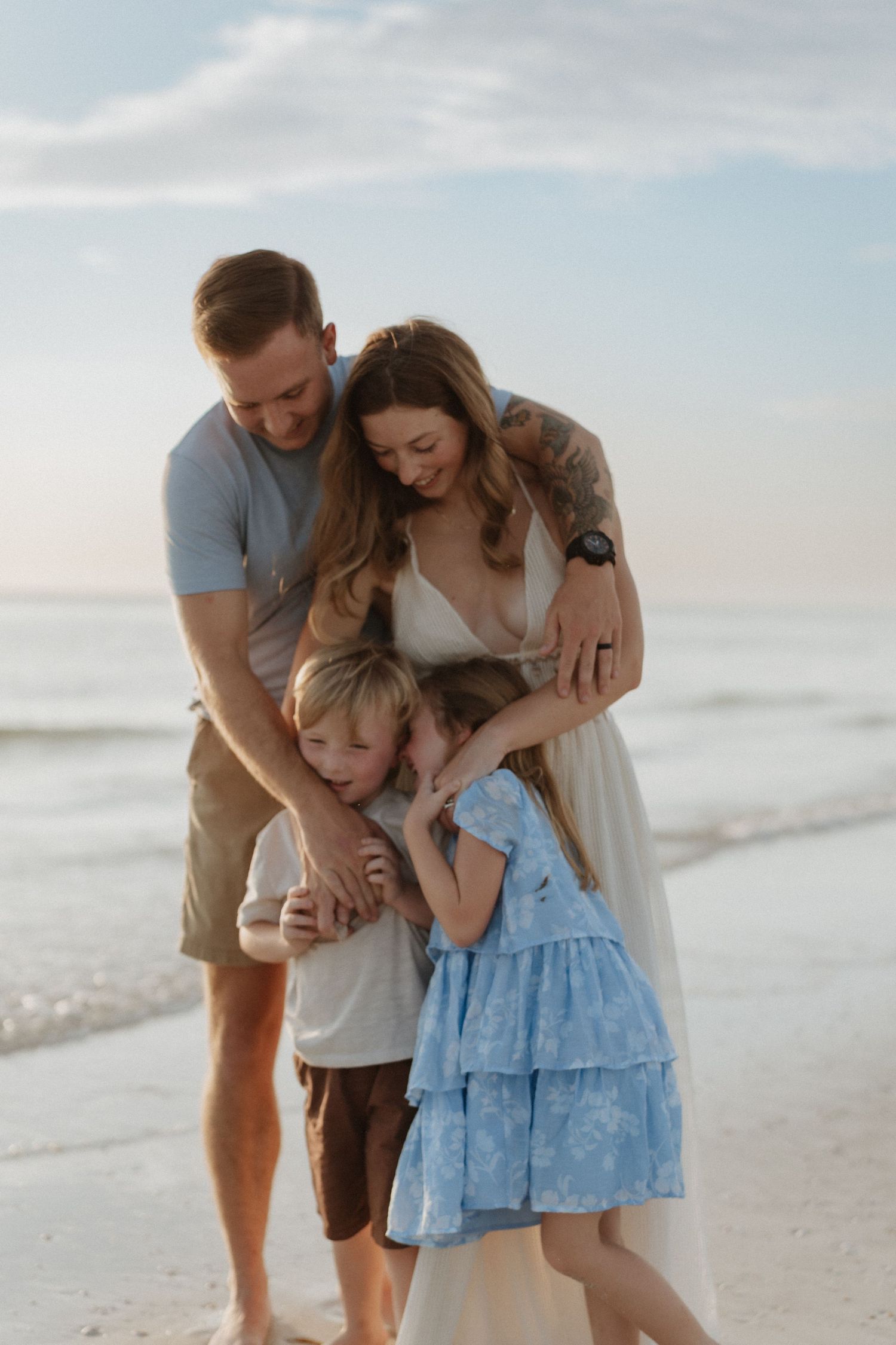 Family of four sharing a warm embrace on a sandy beach at sunset, with two children hugging their parents.
