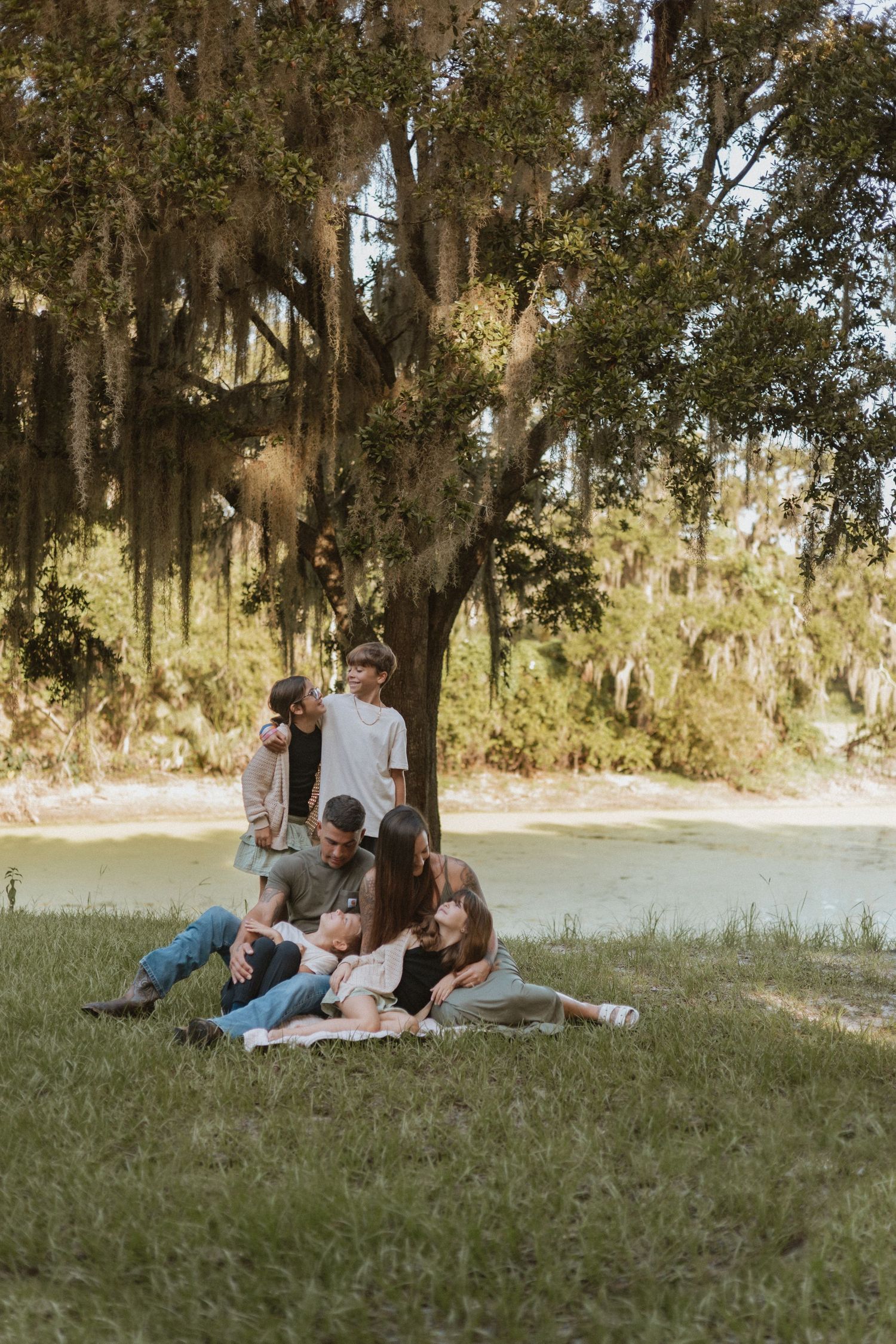 Family lounging on grass under Spanish moss-draped trees during outdoor photoshoot.
