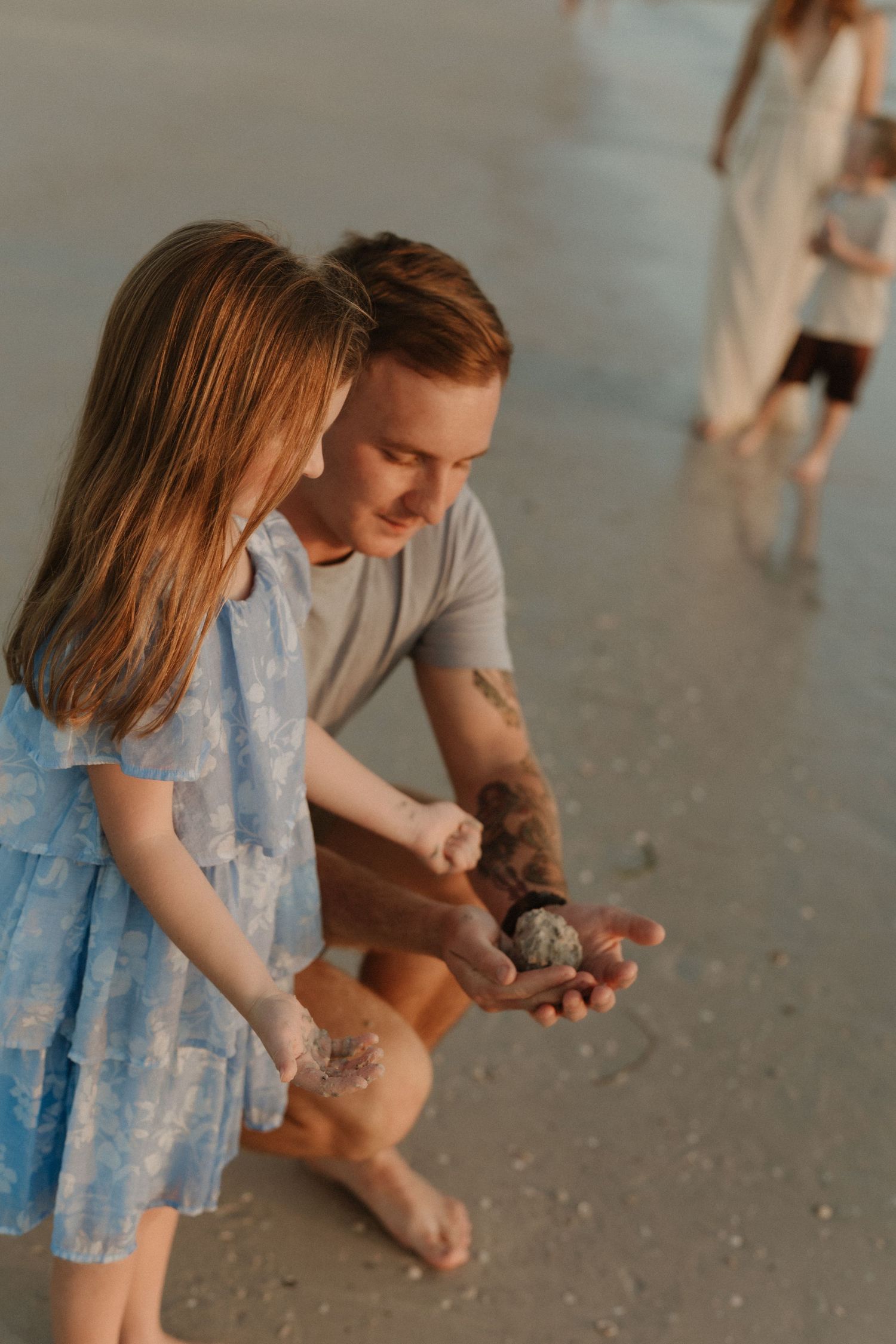 A man and young girl examine a shell on a sandy beach at golden hour, with others visible in the background.
