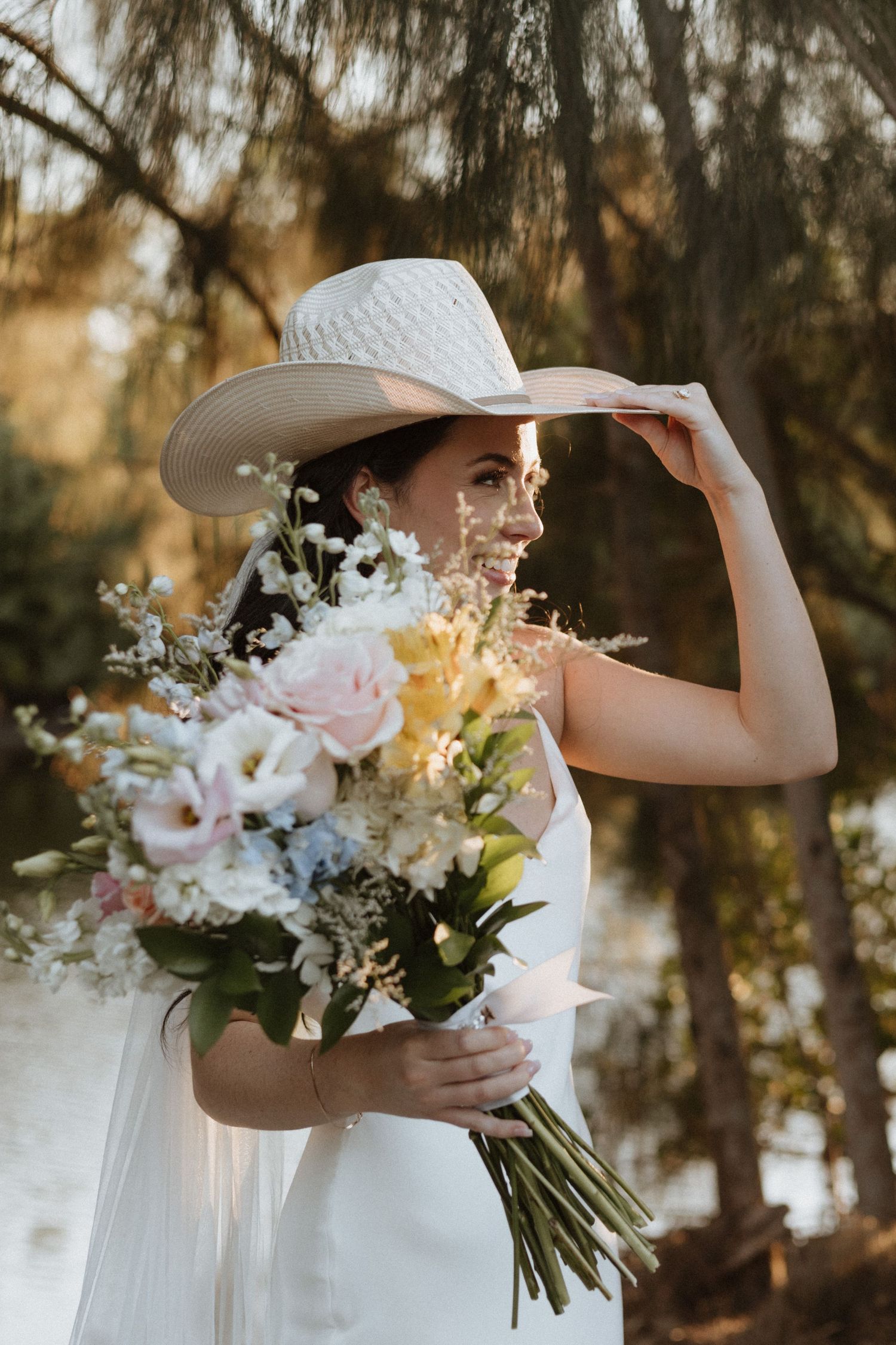 Person in white dress and cowboy hat holding beautiful floral bouquet against natural backdrop.