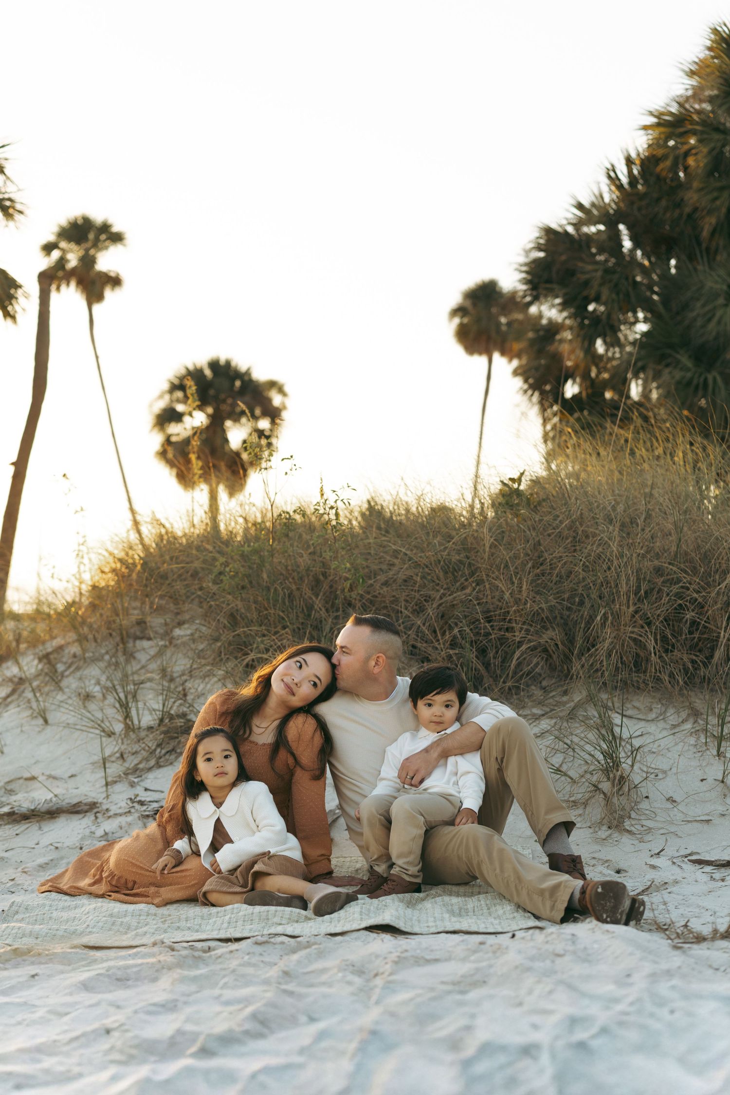 A family relaxes together on a sandy beach at sunset with palm trees silhouetted in the background.