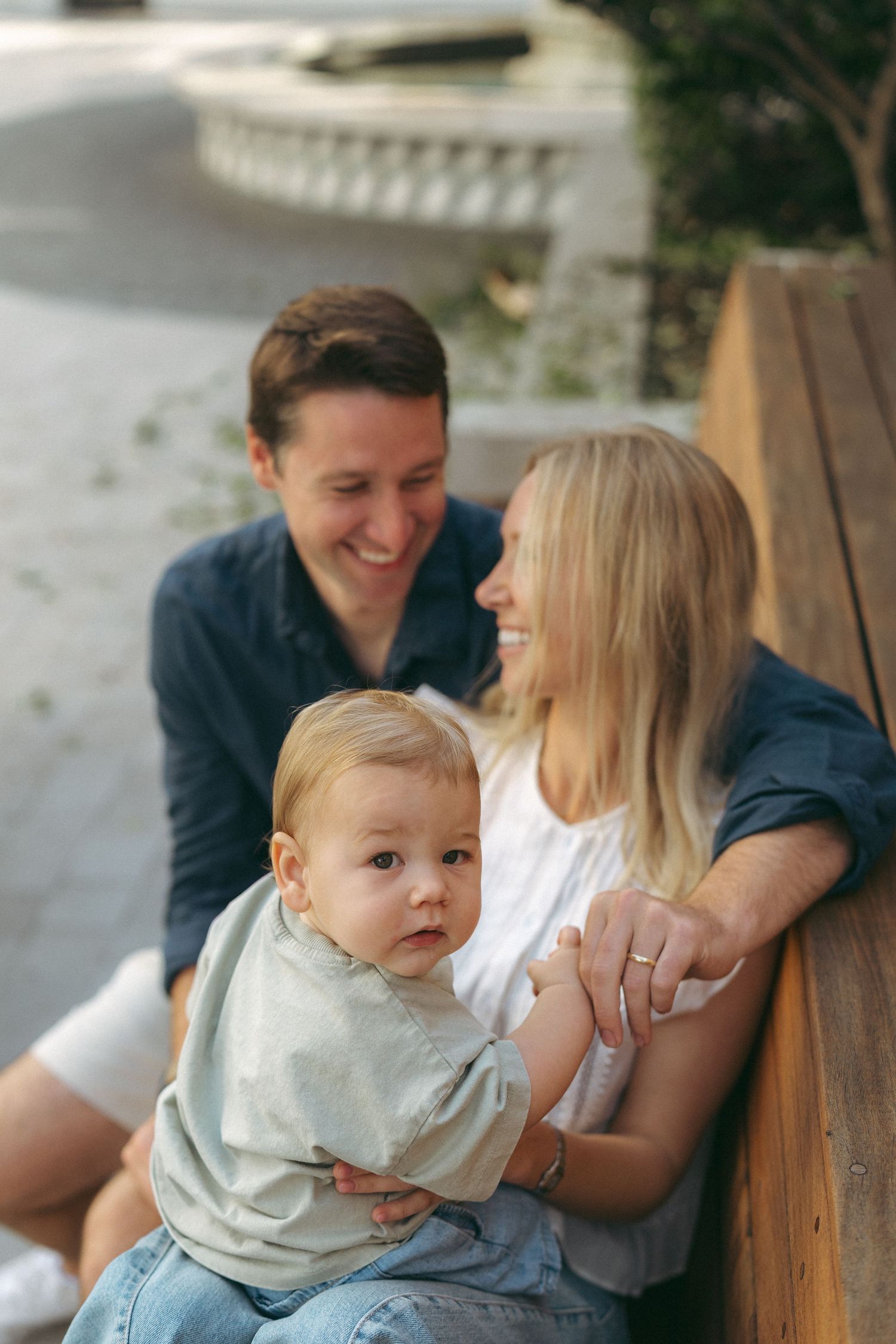 Happy young family with smiling parents and curious baby sitting on wooden bench outdoors in casual clothing.
