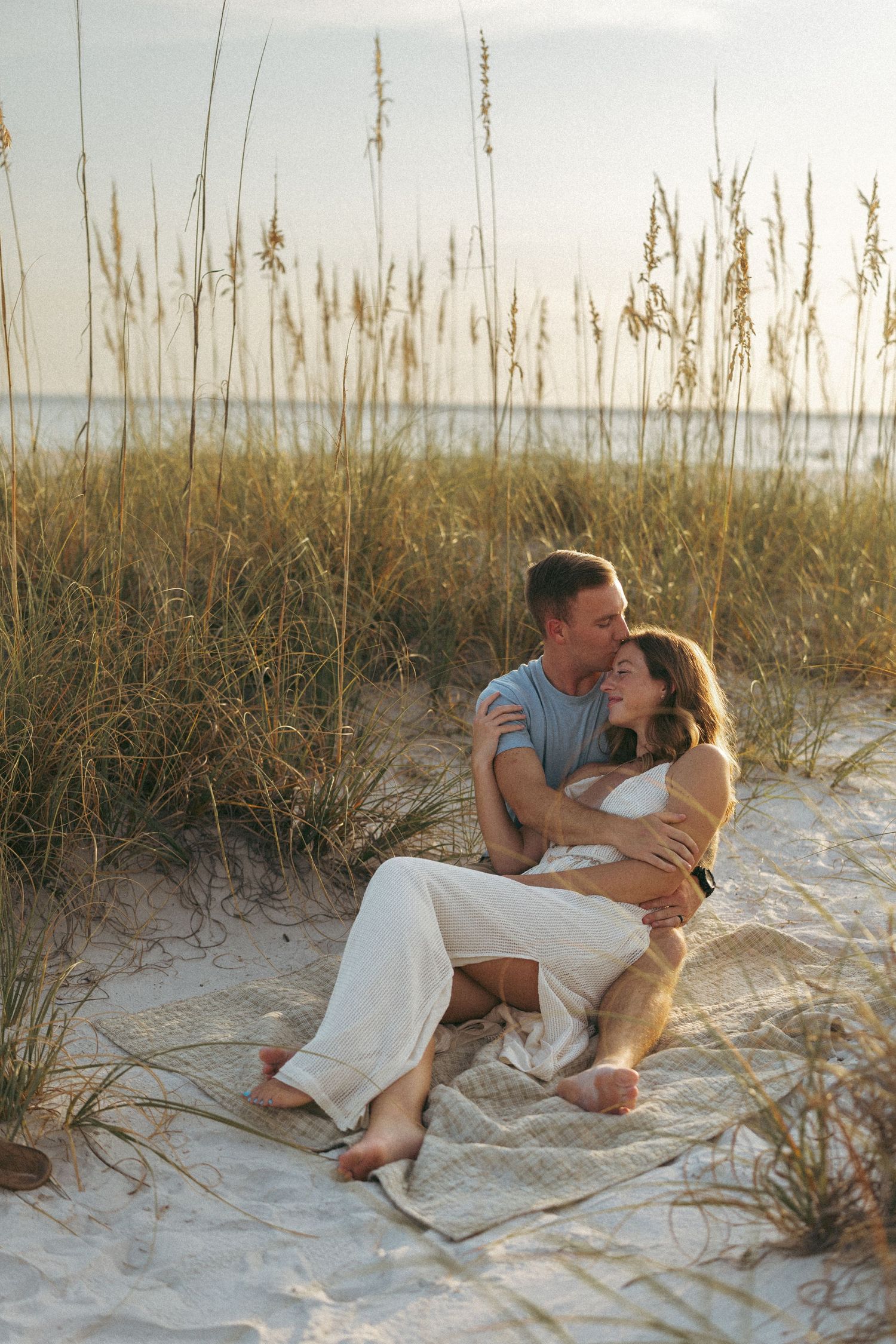 Couple sitting together on sandy beach dunes at sunset, embracing among tall sea oats near the ocean.