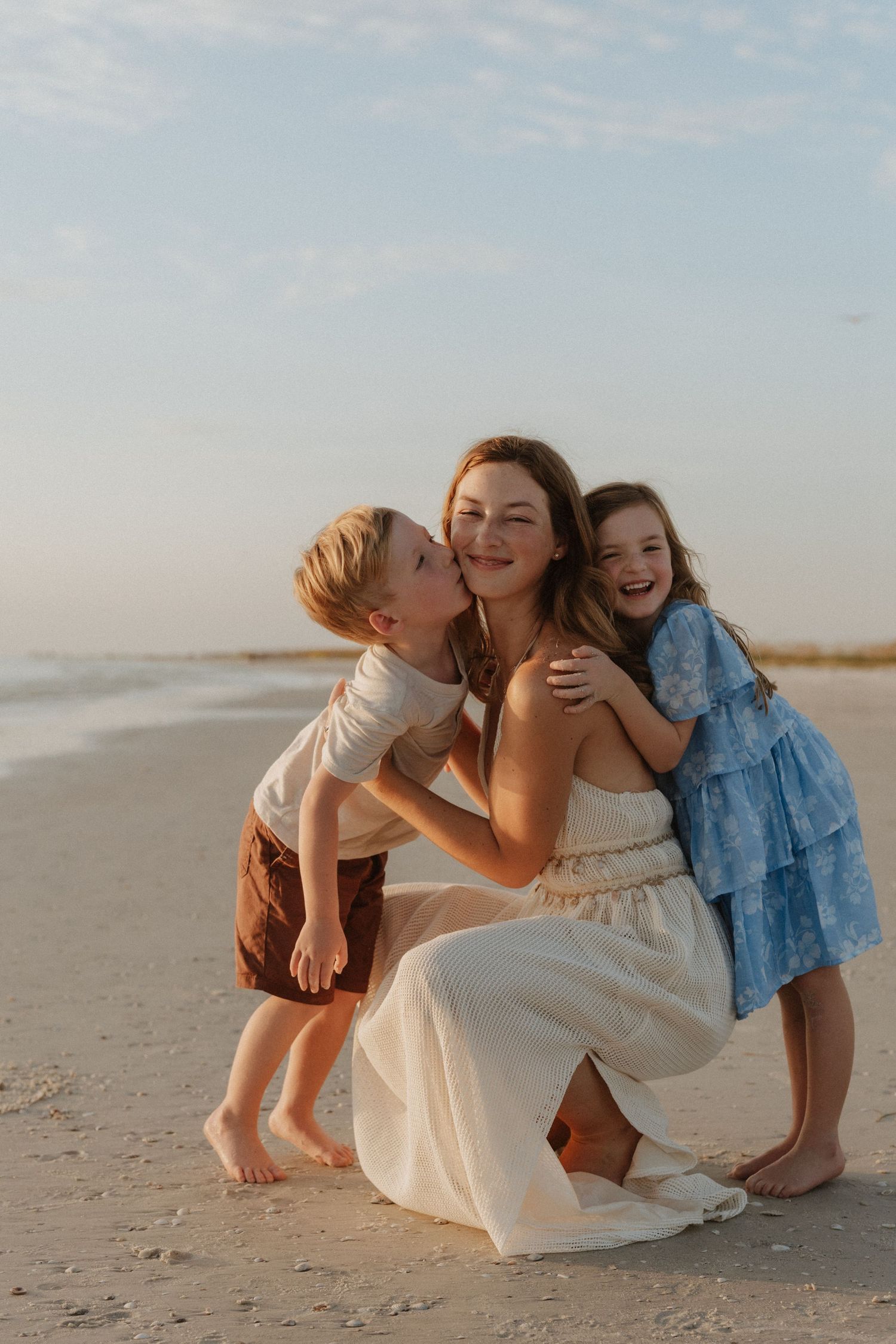 A mother in a white dress crouches on a sandy beach while her young son kisses her cheek and daughter hugs her, smiling.
