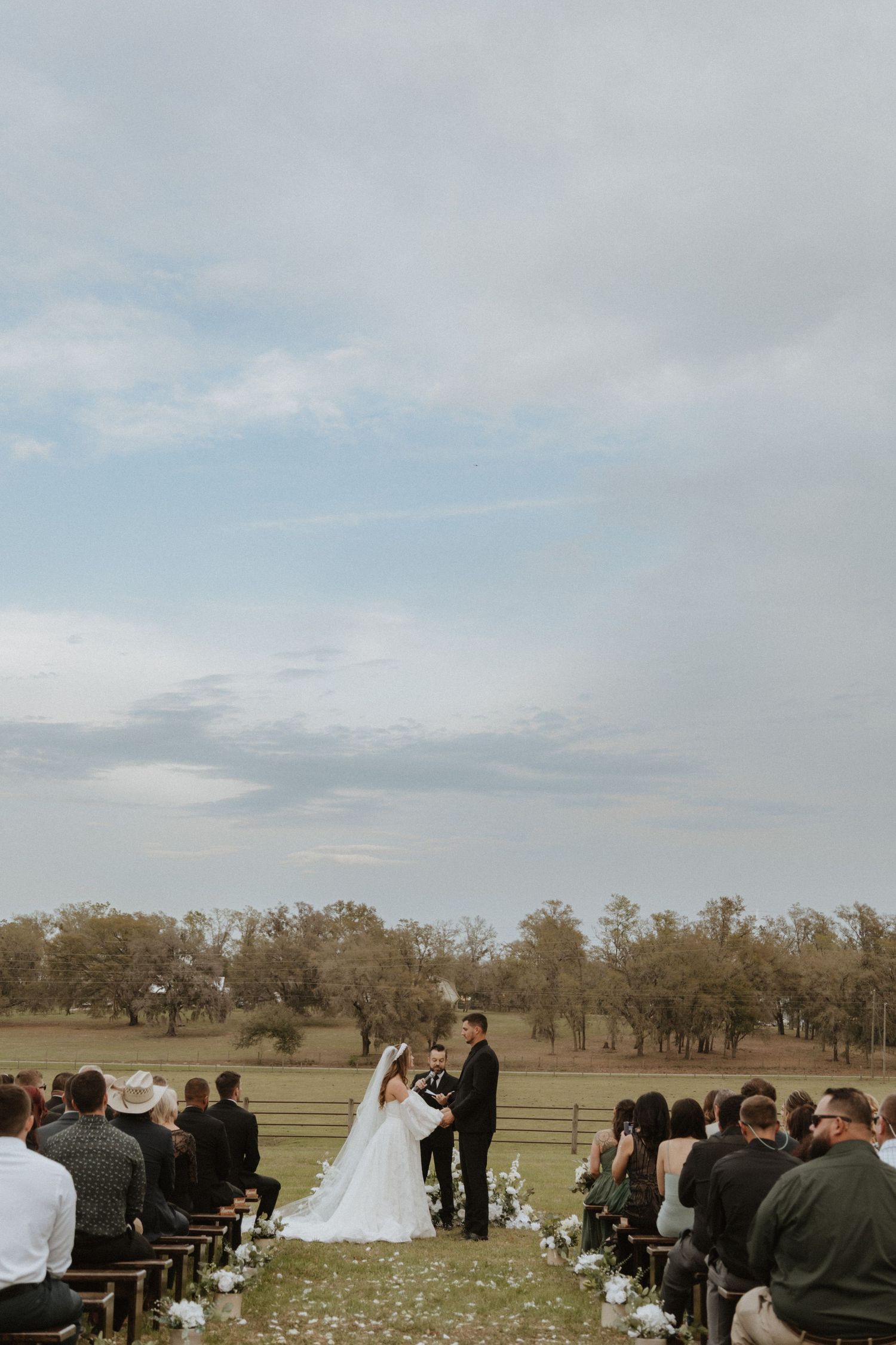 An outdoor wedding ceremony setup with guests seated on white chairs in a field surrounded by trees.
