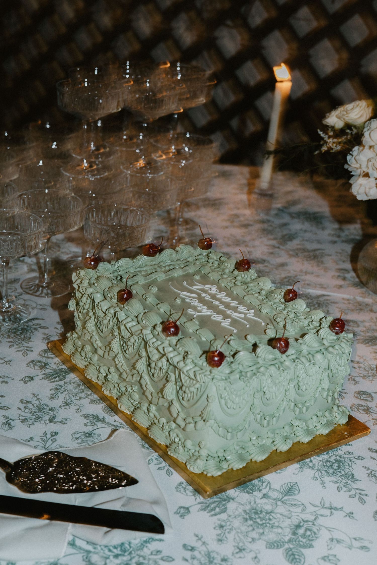 Mint green wedding cake with cherry decorations and champagne glasses in background.