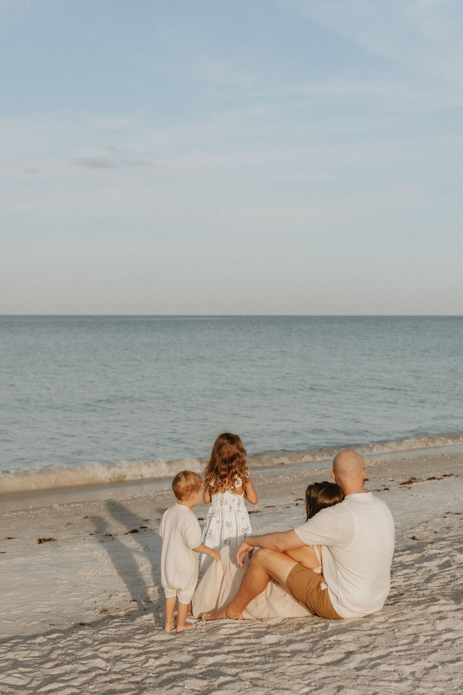 Family of four sitting on a sandy beach at sunset, gazing at calm turquoise ocean waves together.