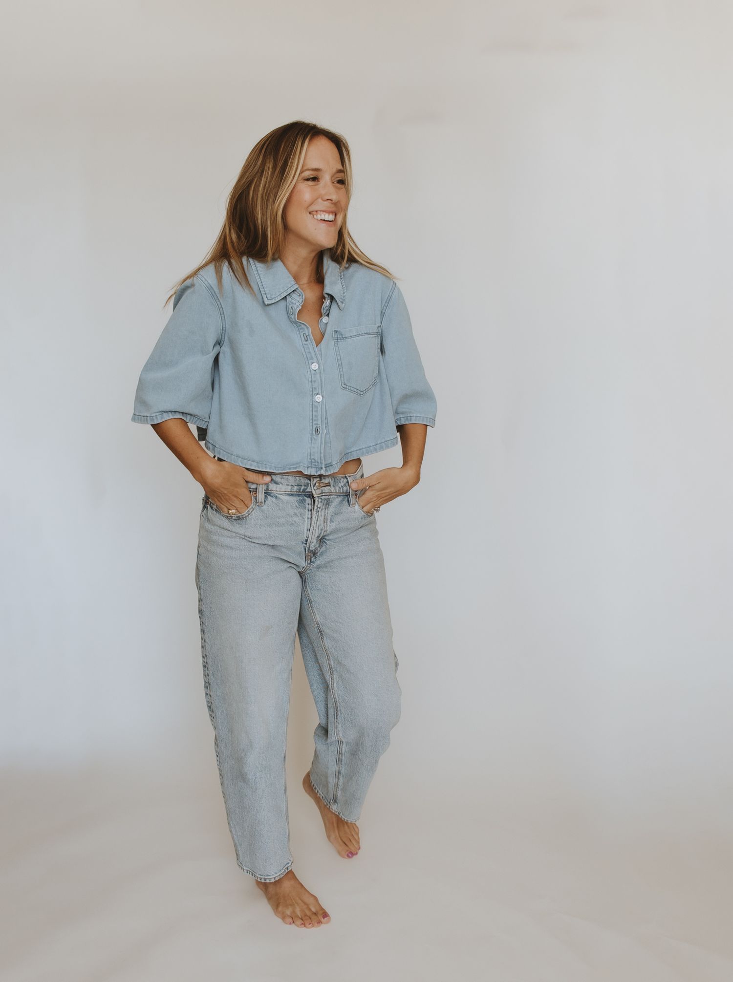 Woman smiling in light wash denim shirt and jeans, standing barefoot against white background.