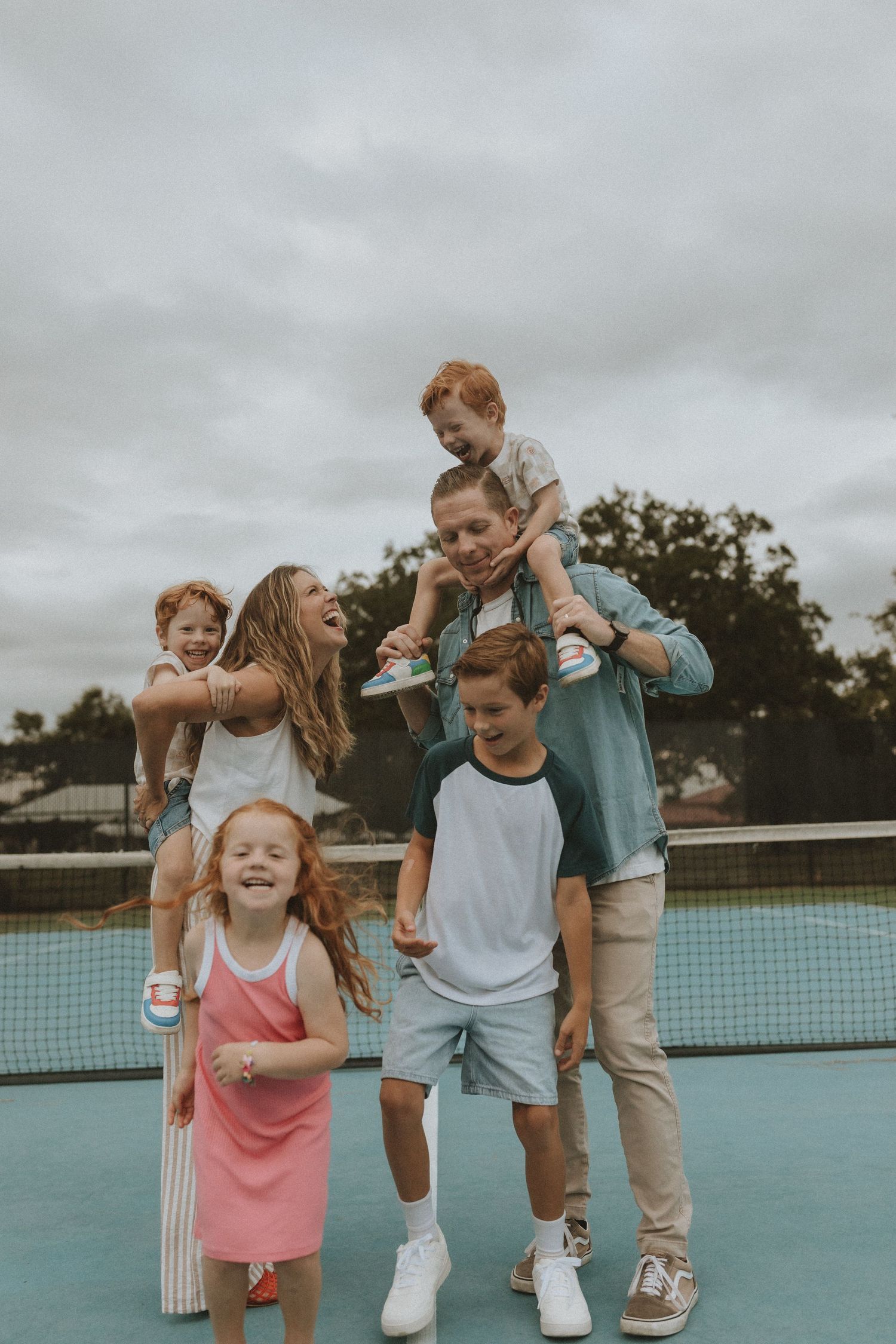 A family shares joyful moments together on a blue tennis court under an overcast sky.