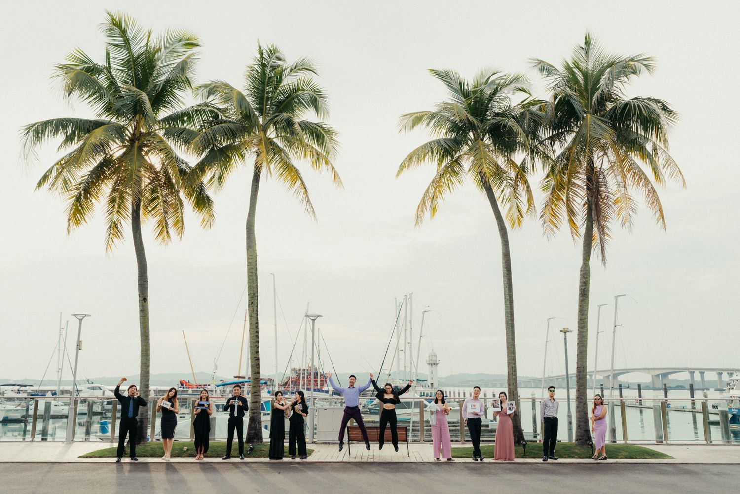 Wedding party celebrates under tall palm trees by a marina, posing joyfully in formal attire on a sunny day.