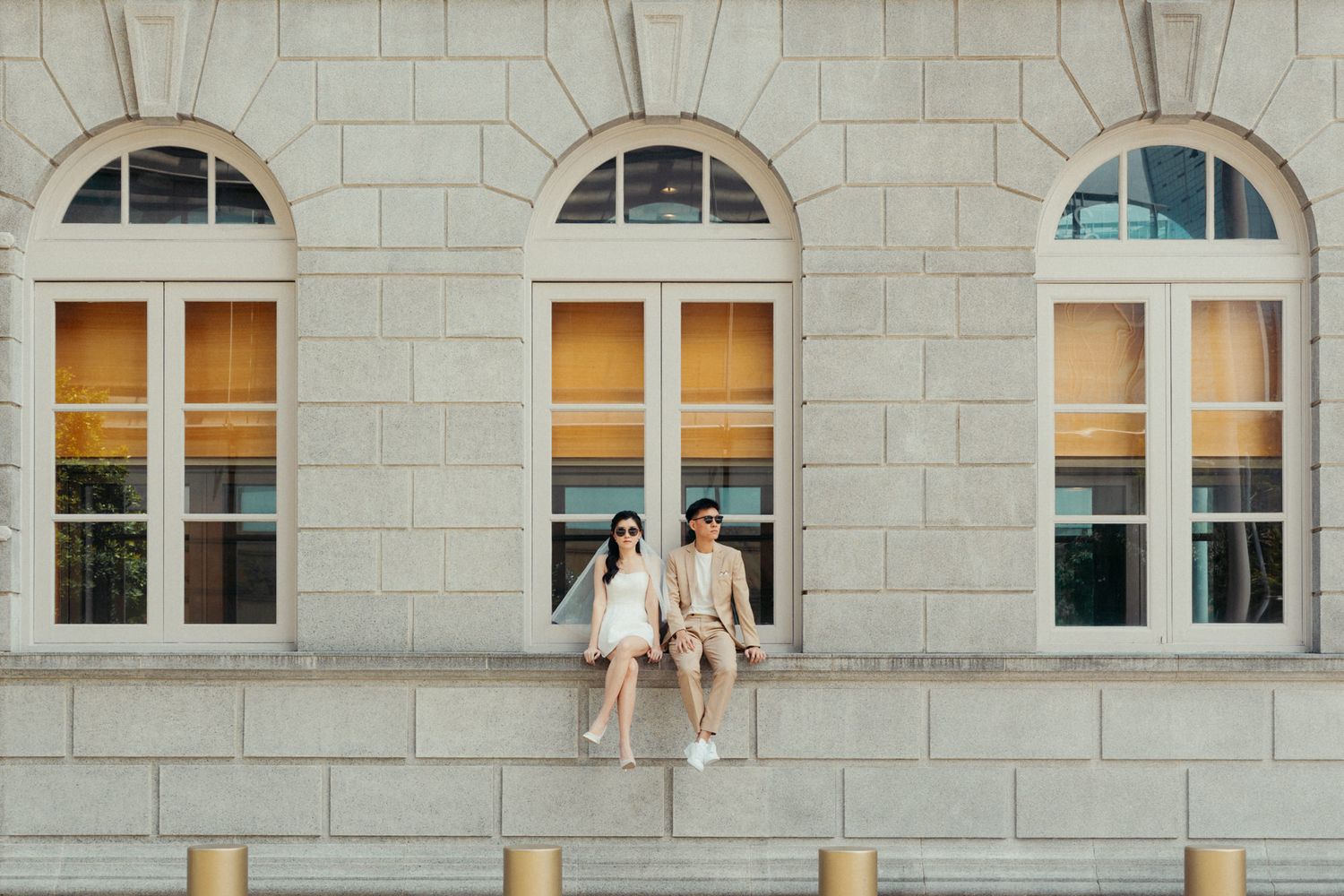 Couple in matching beige outfits sitting on a stone ledge outside a classic building with arched windows.