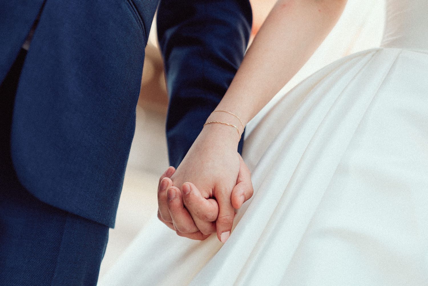 Close-up view of hands holding each other during a wedding ceremony with white dress and navy suit details.
