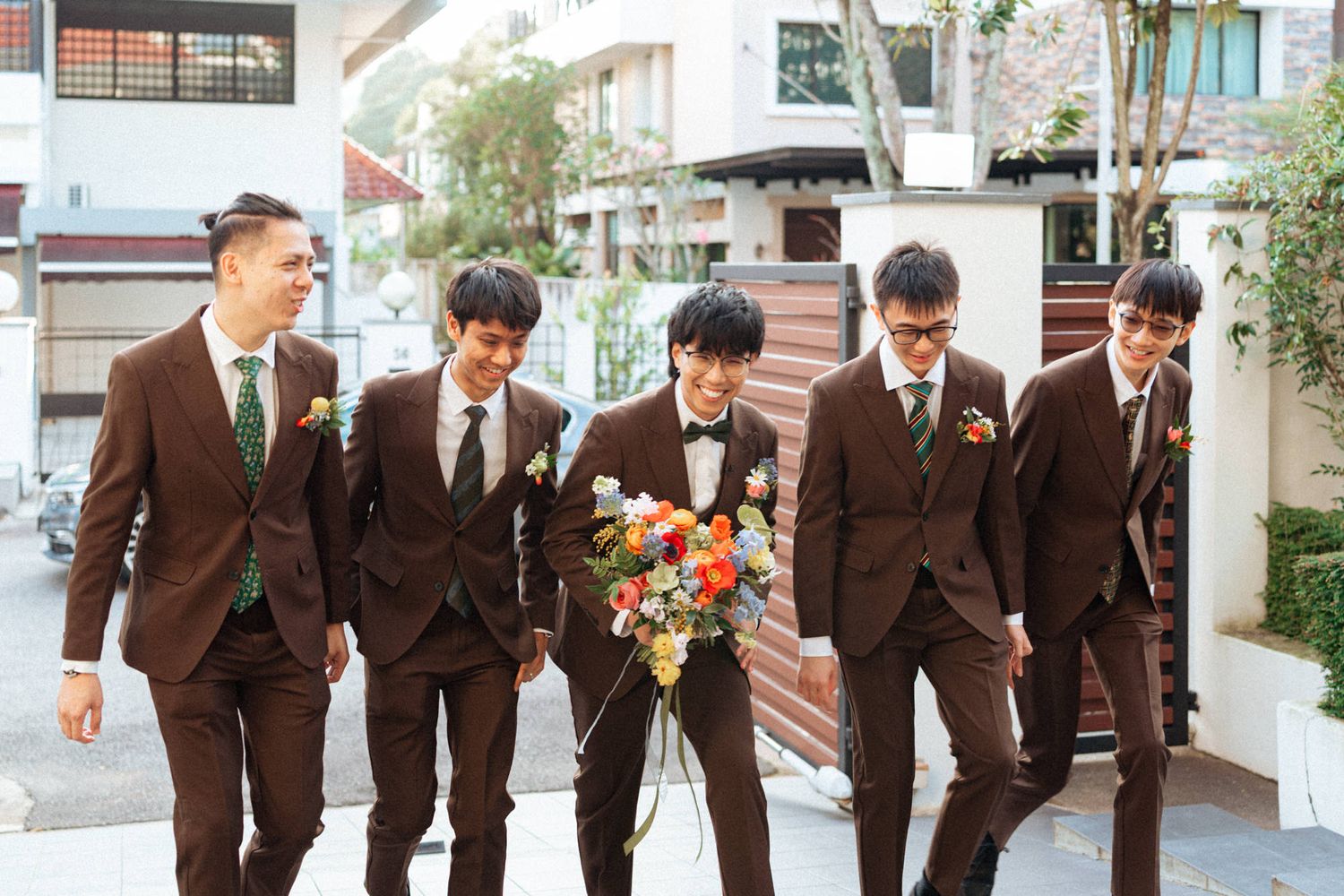 Five groomsmen in brown suits with green ties walk together outdoors, one holding a colorful bridal bouquet.