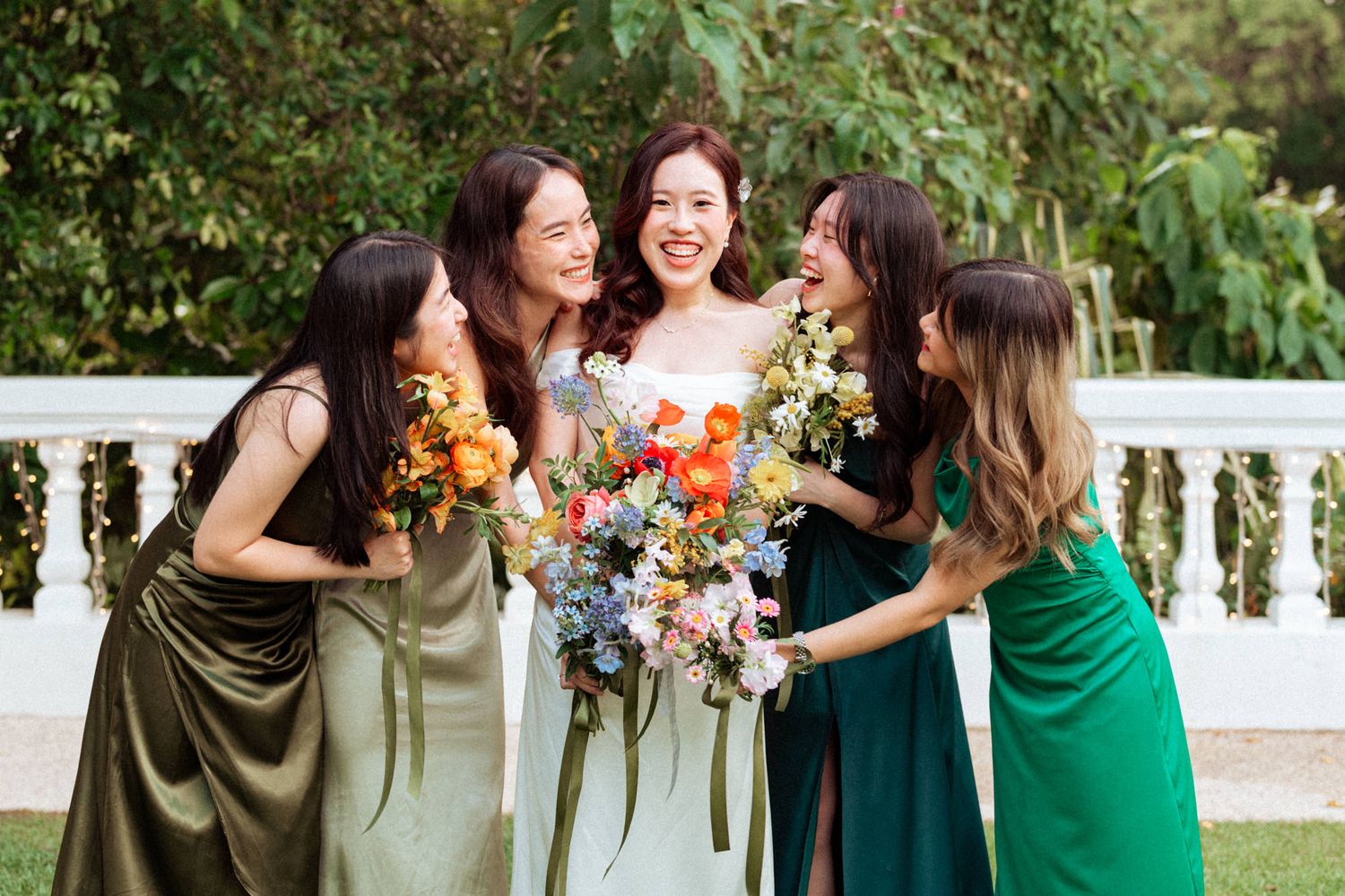 Bride in white gown laughing with four bridesmaids in green dresses holding colorful floral bouquet outdoors.