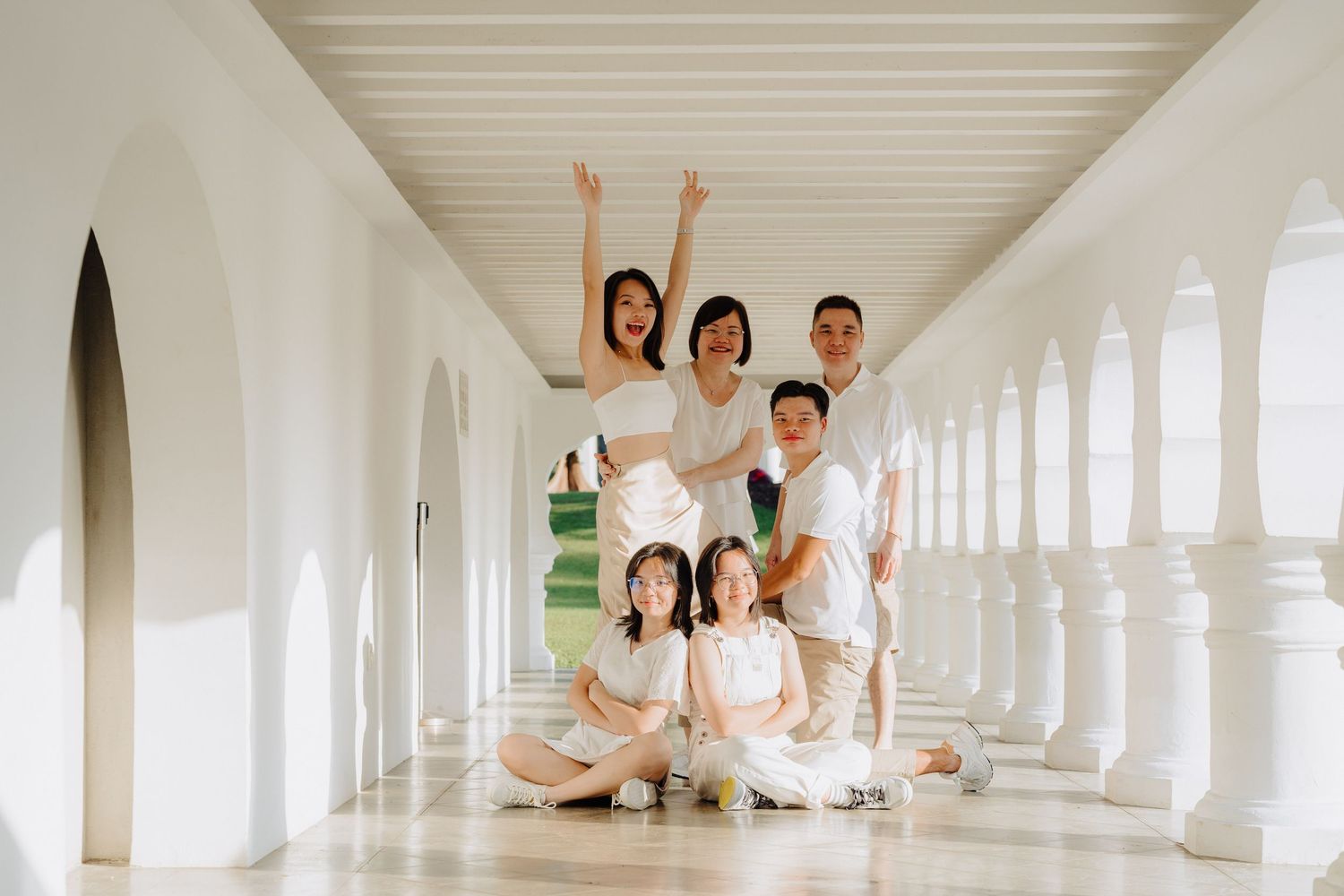 A group in white clothing sits and poses in a bright corridor with white columns and natural lighting.