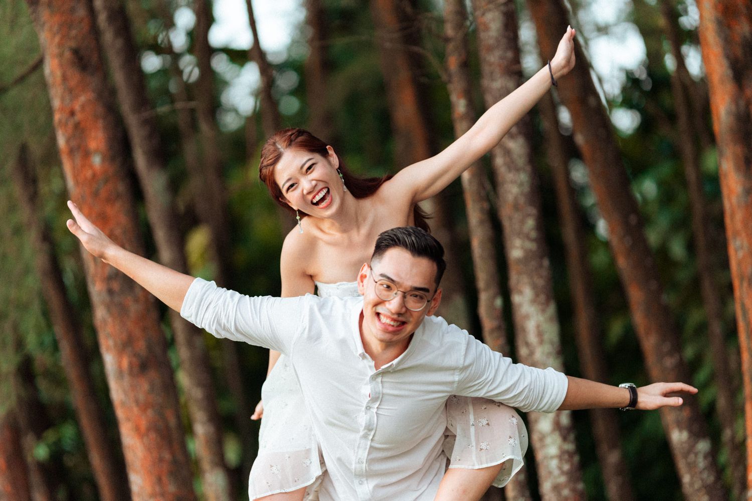 Couple spreads their arms playfully while posing between pine trees.