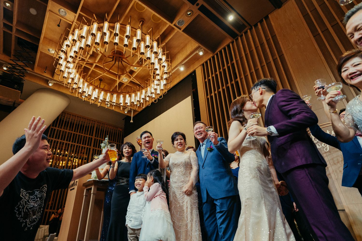 Wedding party celebrating joyfully under elegant chandelier, bride in white gown, groom in blue suit, guests toasting with drinks.