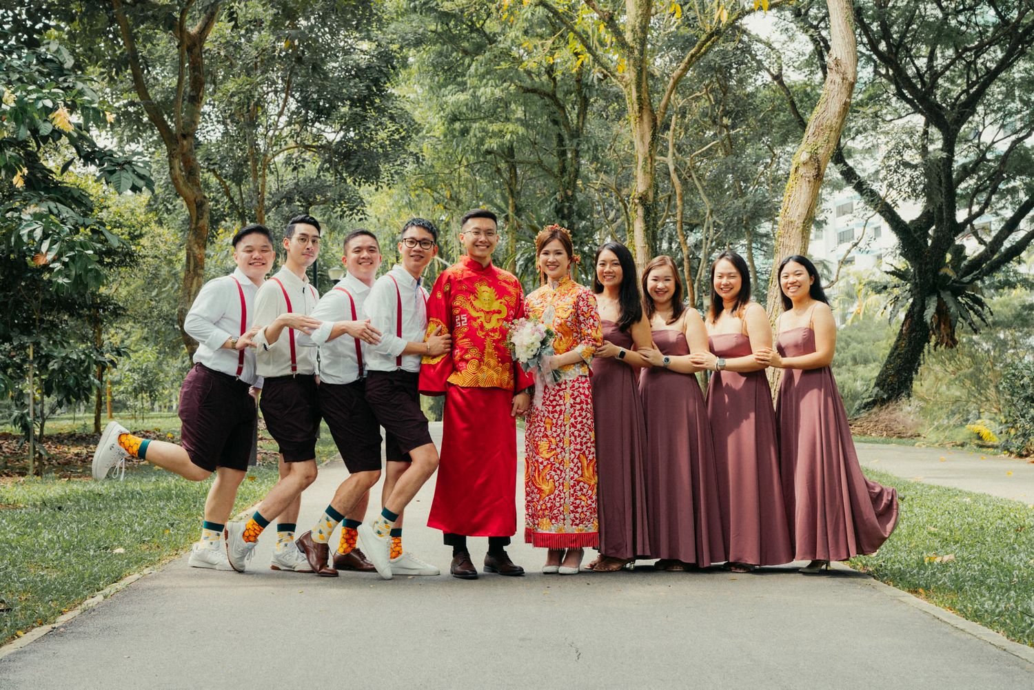 Chinese wedding group photo with bride in red qipao, bridesmaids in mauve gowns, and groomsmen outdoors.