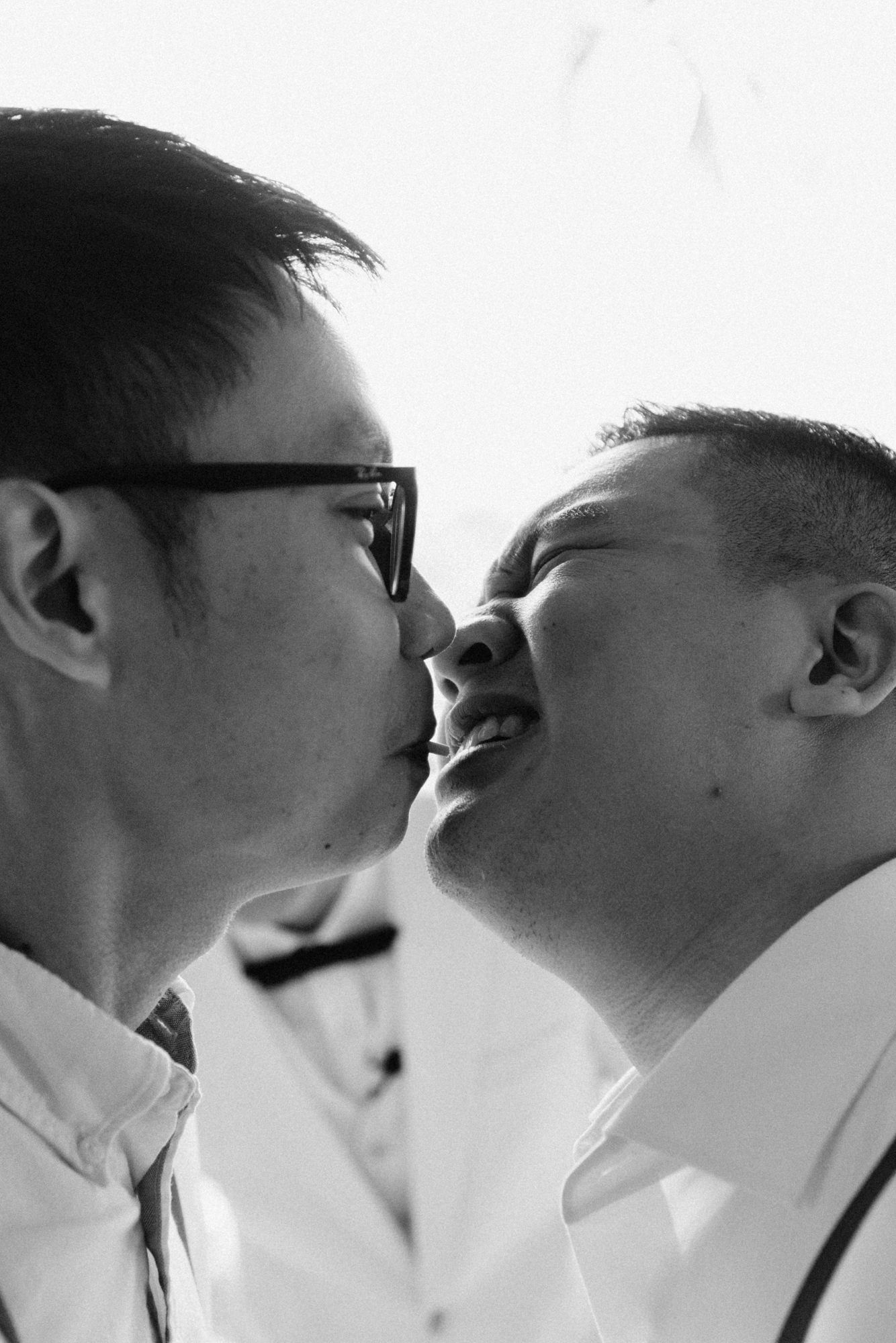 Black and white photo of two men sharing a kiss, one wearing glasses, in formal white shirts.