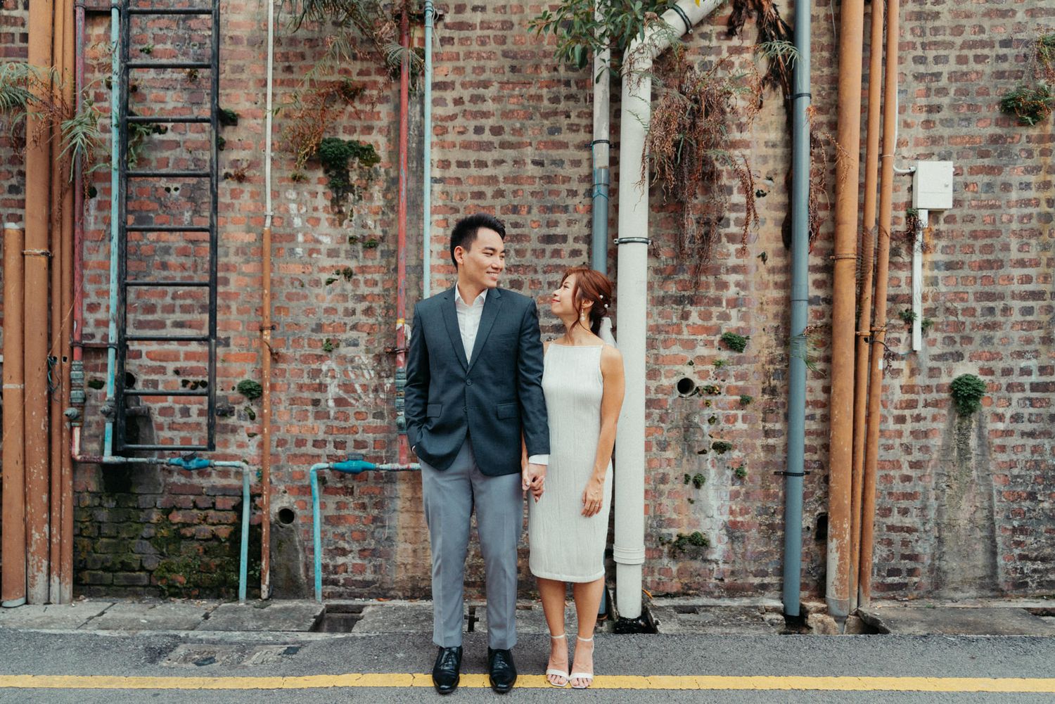 Couple standing together in front of a rustic brick wall, man in suit and woman in white dress, urban outdoor setting.
