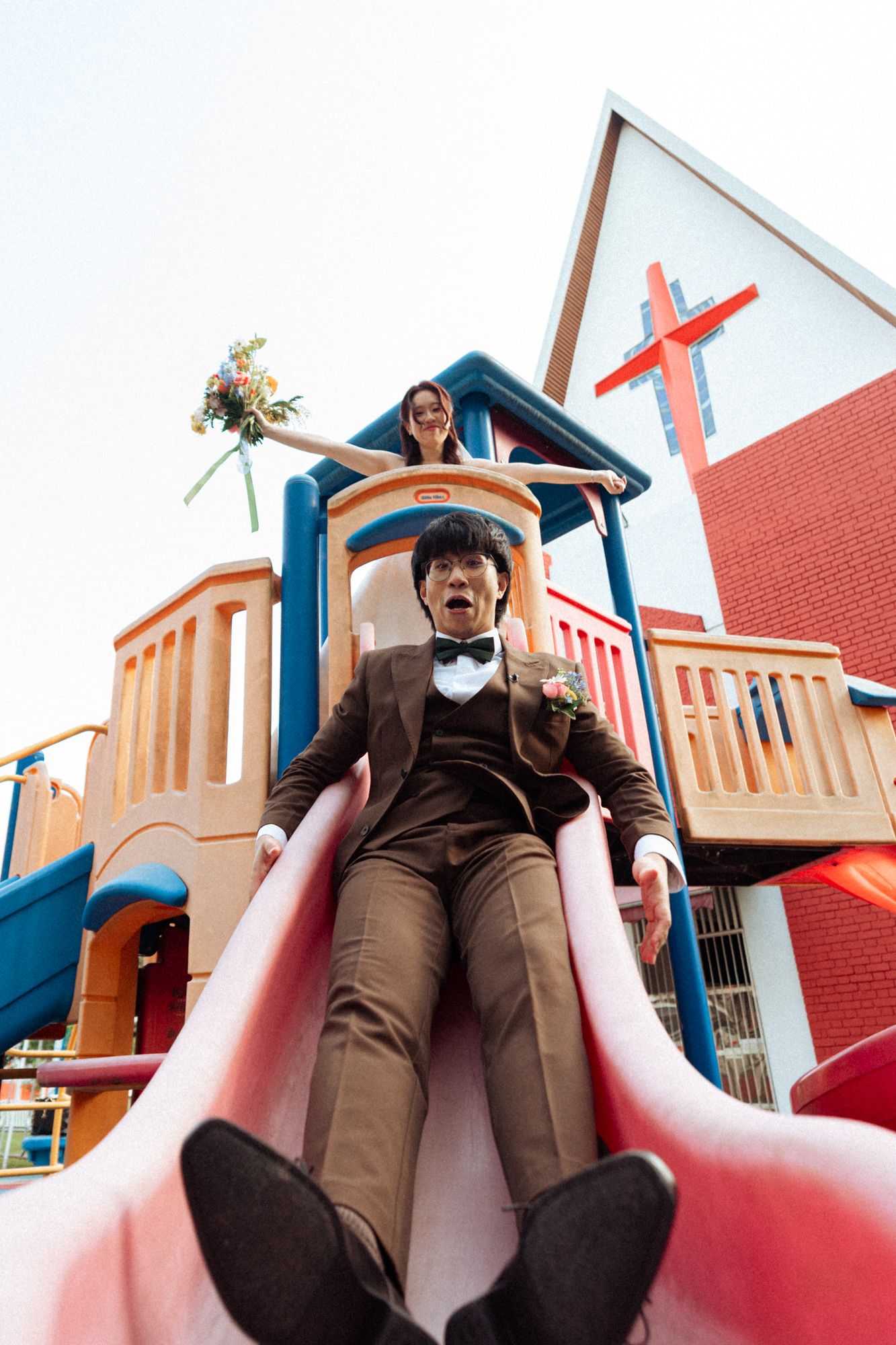 Groom in brown suit slides down playground slide with bride holding bouquet above near church cross.