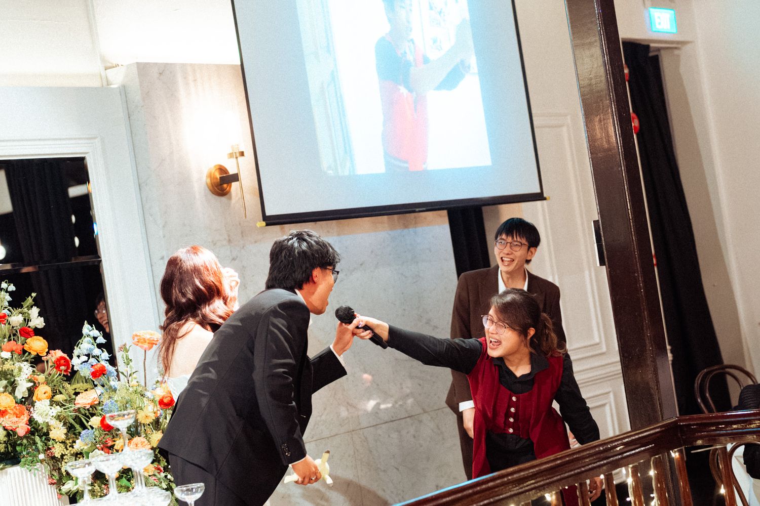 A man holds a microphone toward a woman at an elegant indoor event with floral arrangements and a projection screen.