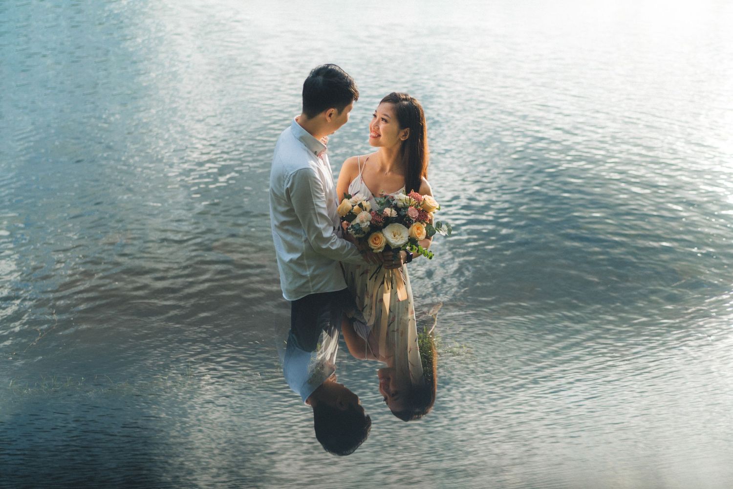 A couple shares a romantic moment while standing in shallow ocean water with a floral bouquet at sunset.