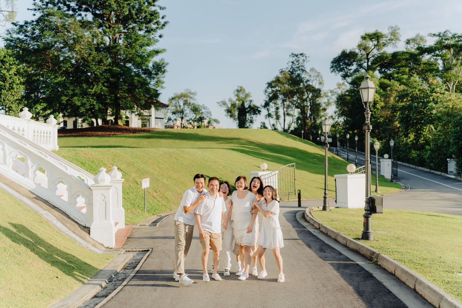Group in white outfits poses together on a grassy slope with trees and architecture in the background.