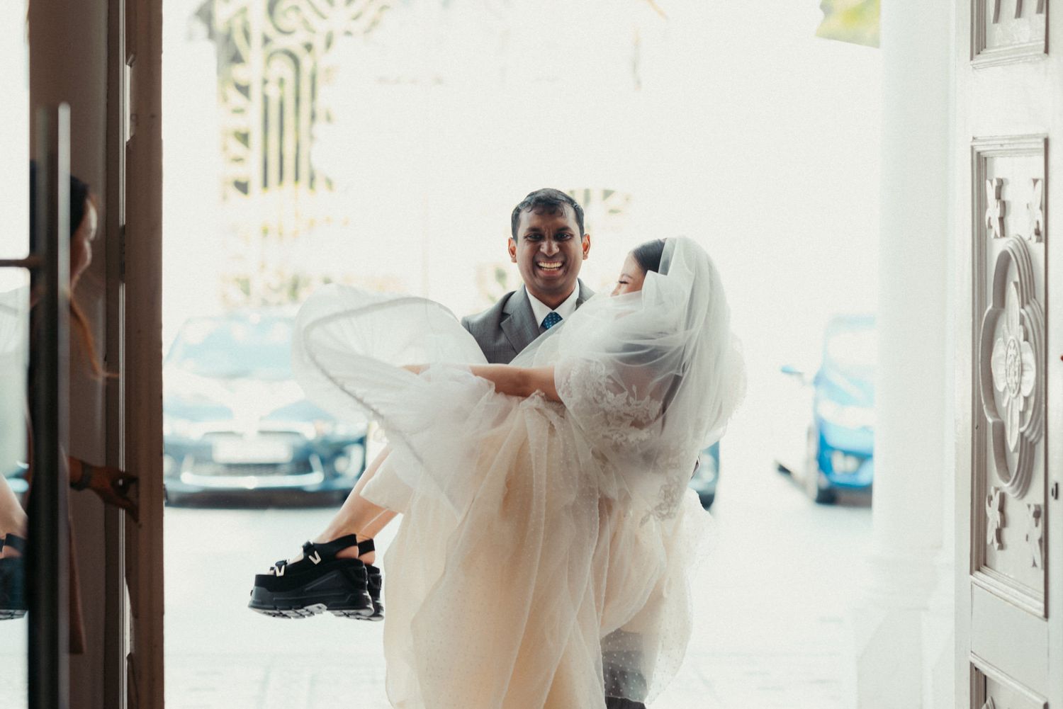 Groom carrying bride in white wedding dress through church doorway on their wedding day.