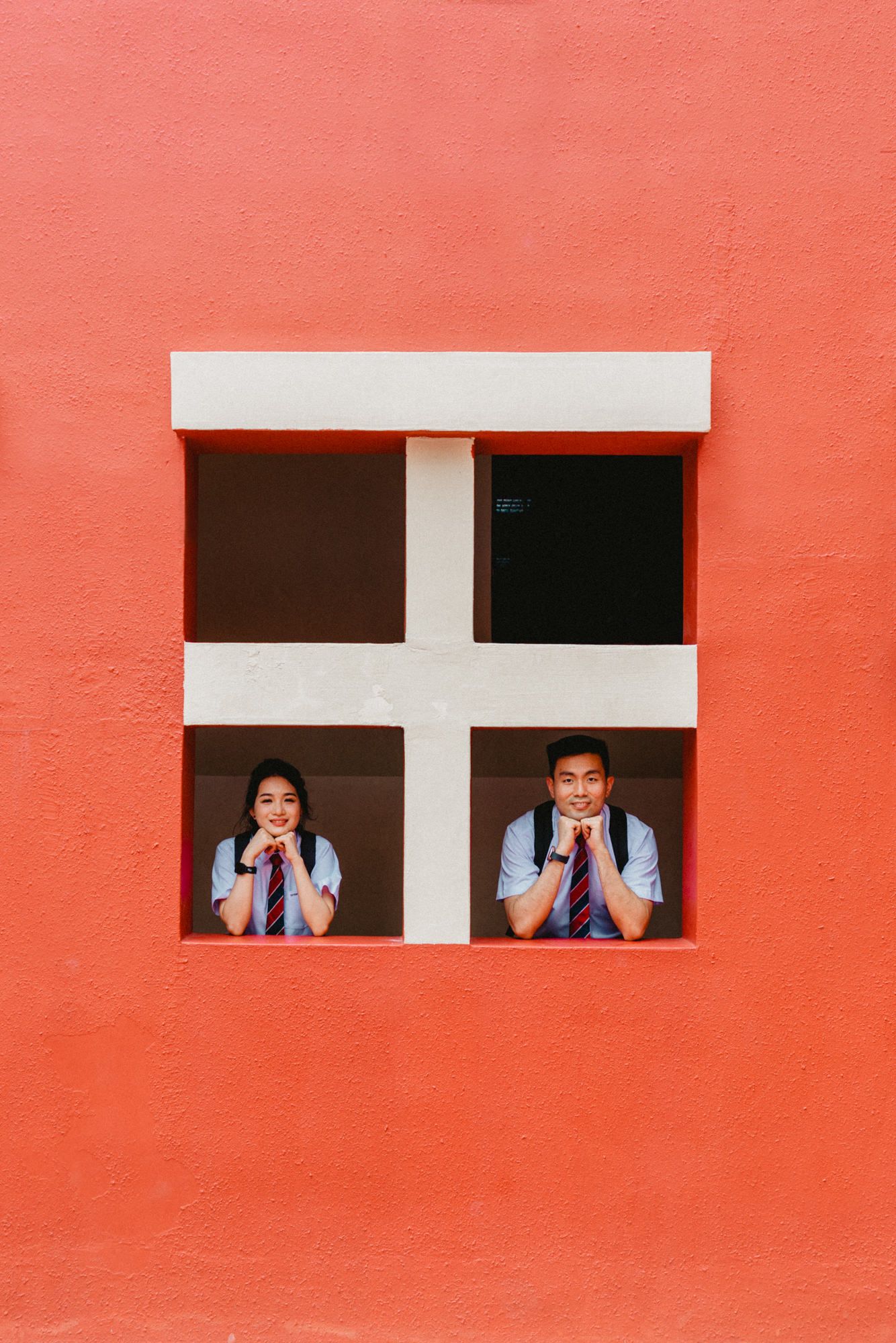 Two students peek through a cross-shaped window frame in a coral-colored wall.