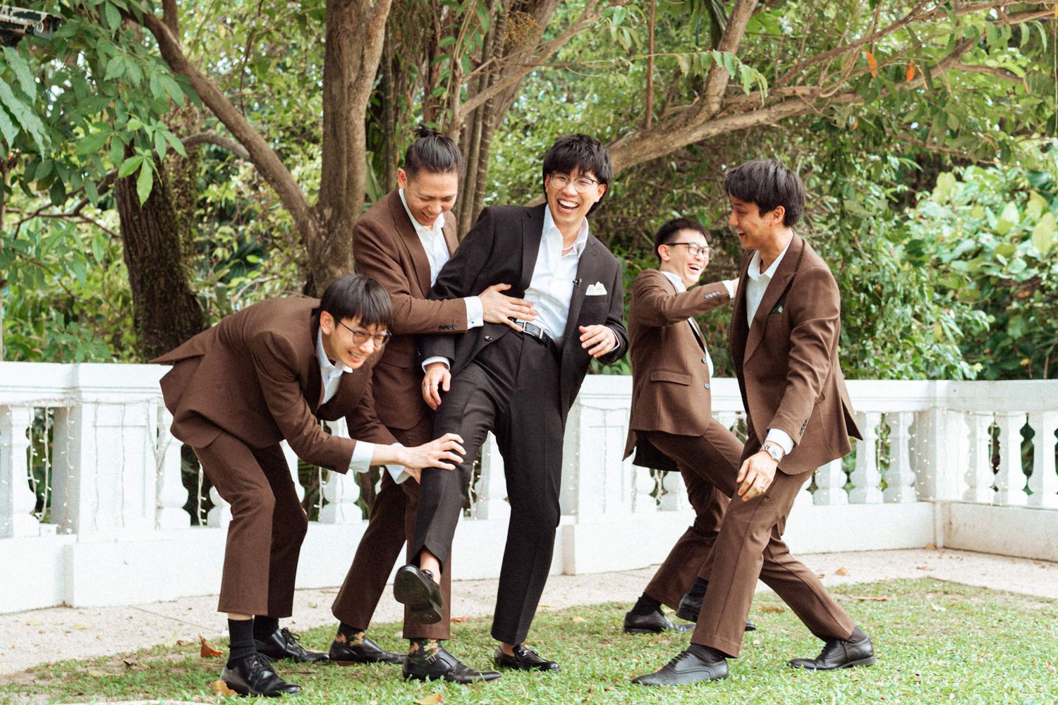 Five young men in matching brown suits pose playfully outdoors near a tree and white railing.